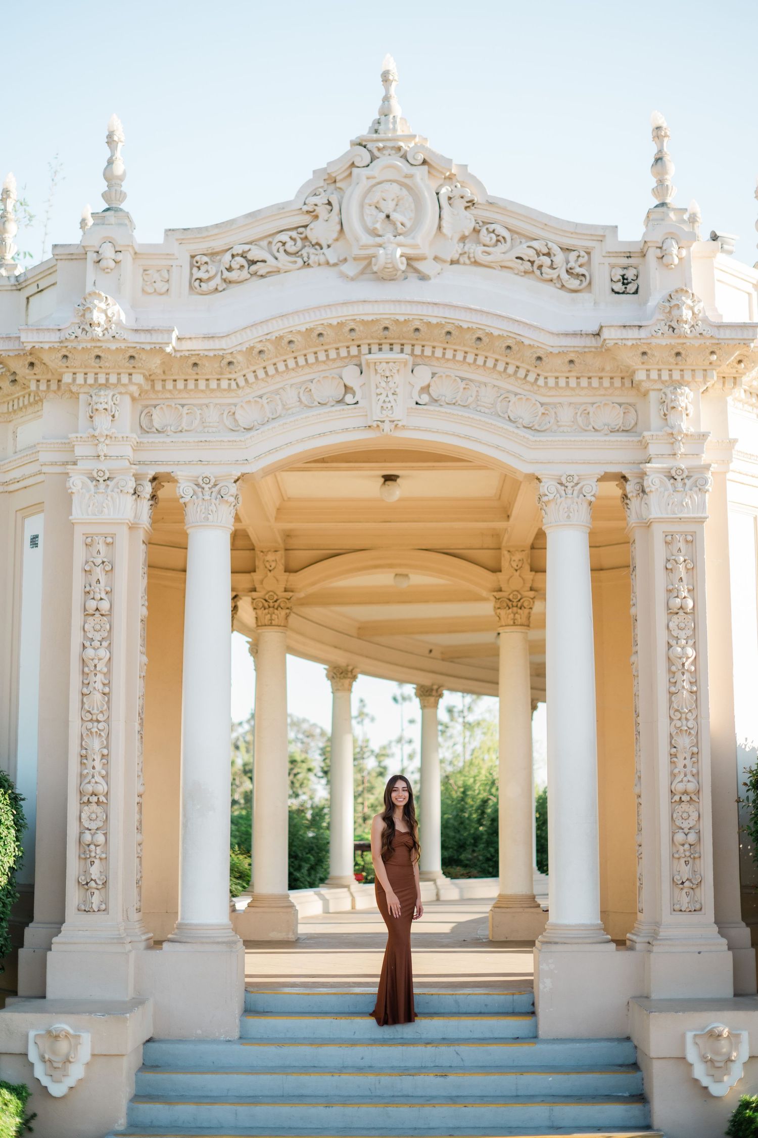 Woman in brown gown poses at ornate white Baroque colonnade with decorative columns and arched entrance.