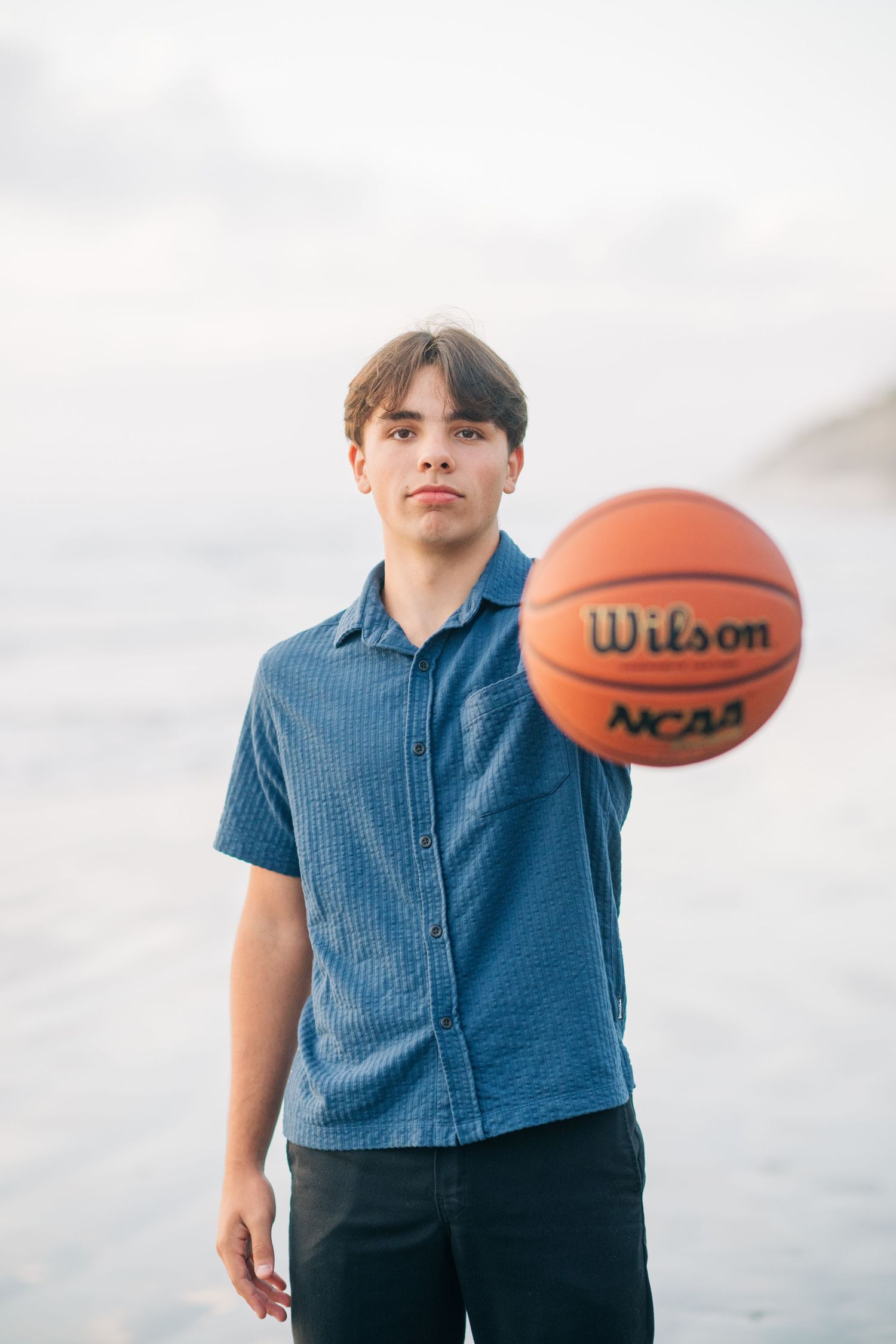 Teen boy in blue shirt holding a Wilson NCAA basketball on a beach, looking confidently at the camera.