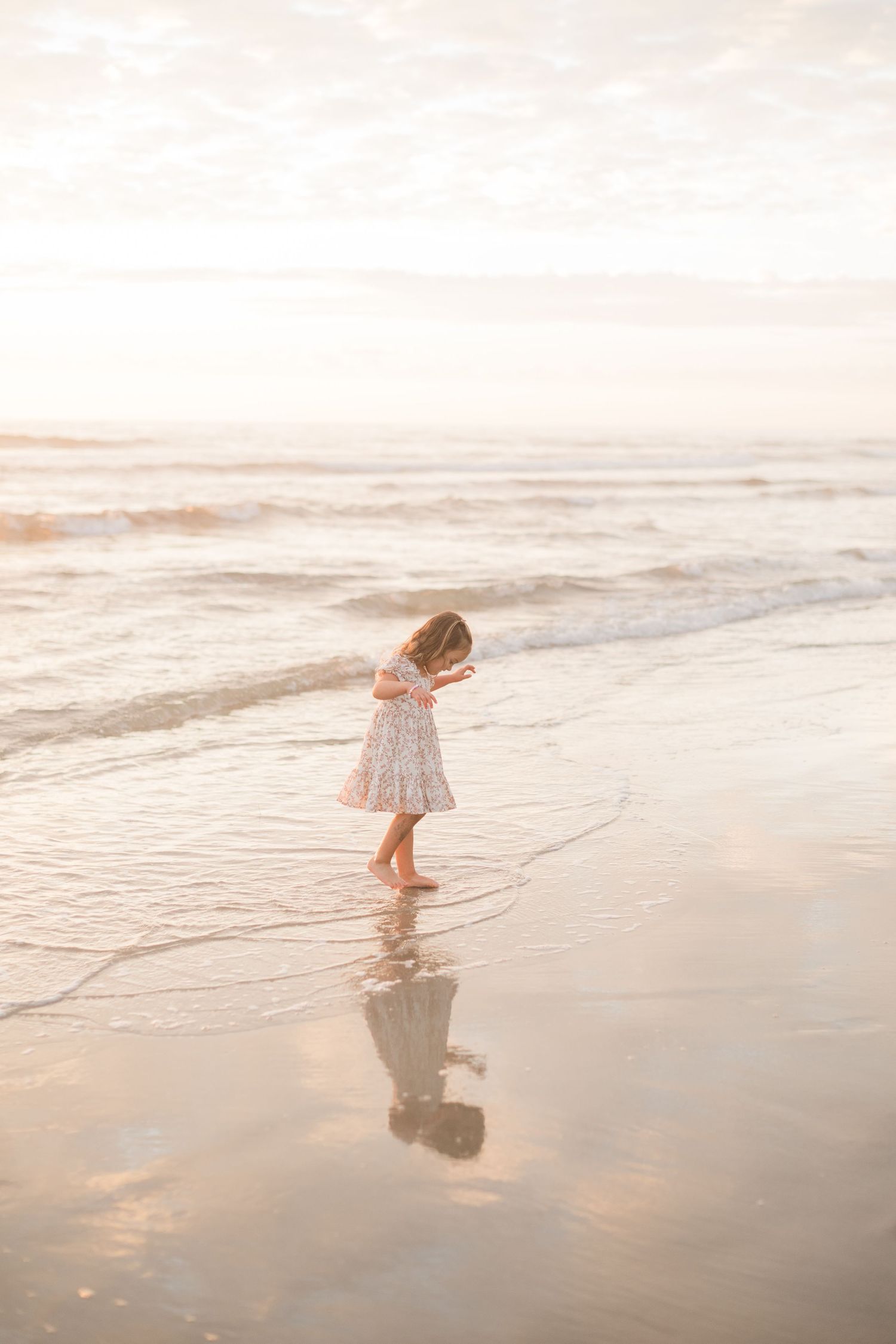 Young girl in a floral dress walking barefoot along the shoreline at sunset, reflected in wet sand.