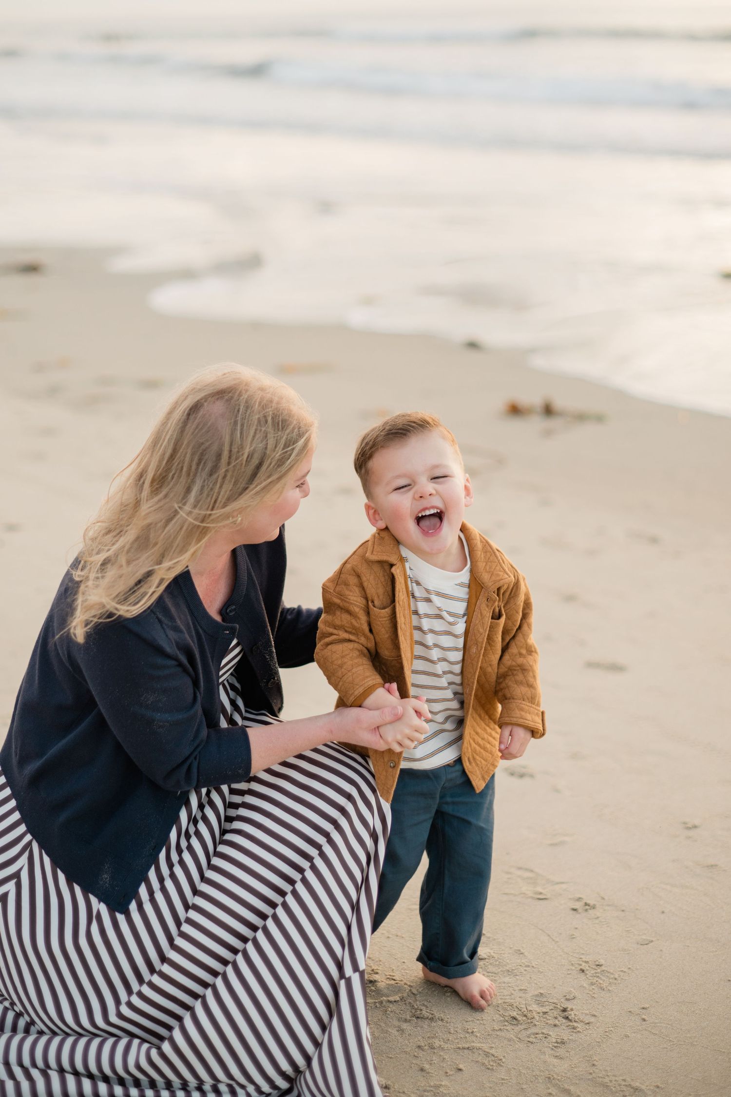 A laughing toddler boy holds his smiling blonde mother's hands on a sandy beach at sunset.