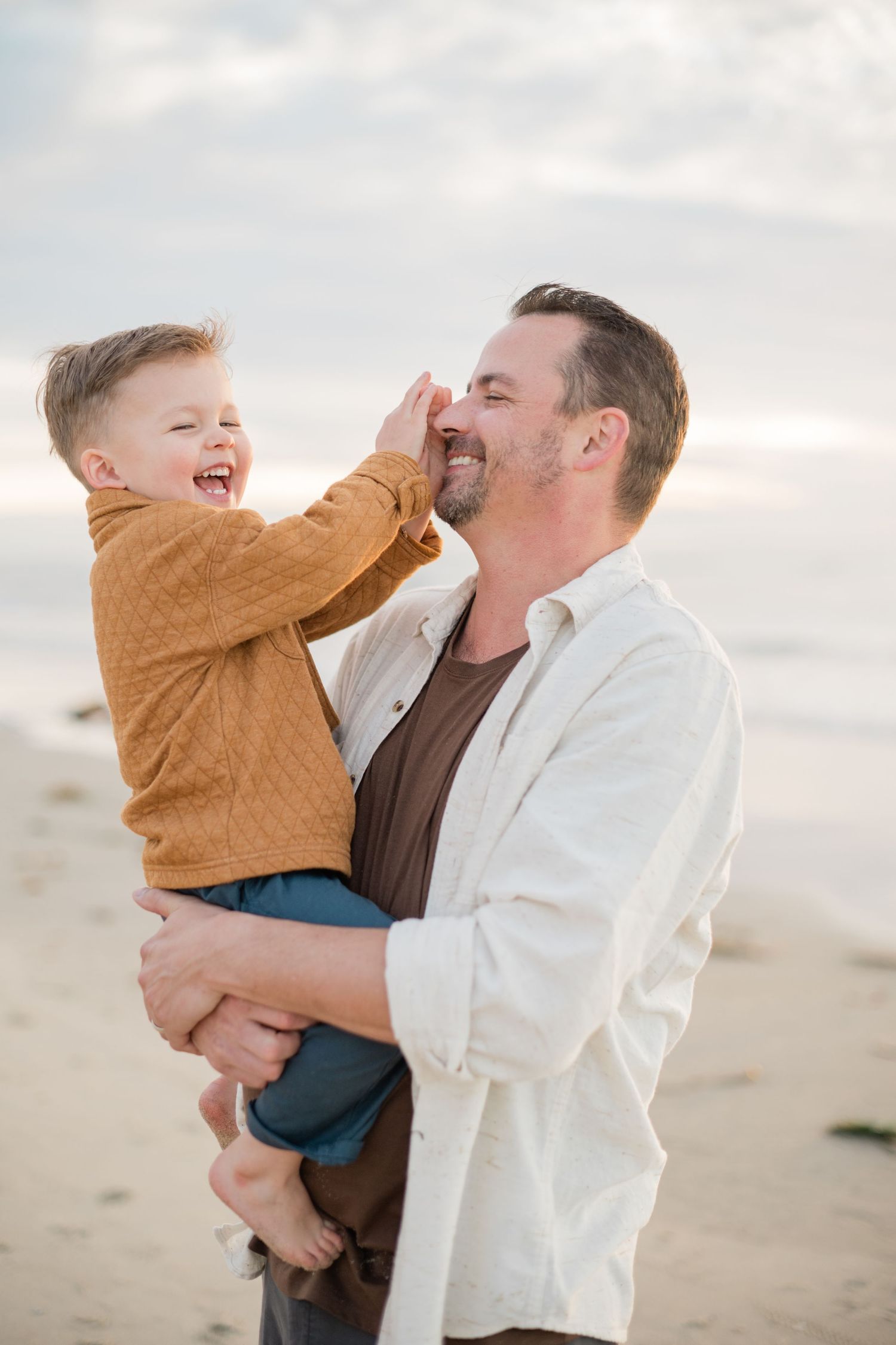 A father holds his laughing toddler son on a beach, sharing a joyful moment as the boy touches his dad's face.