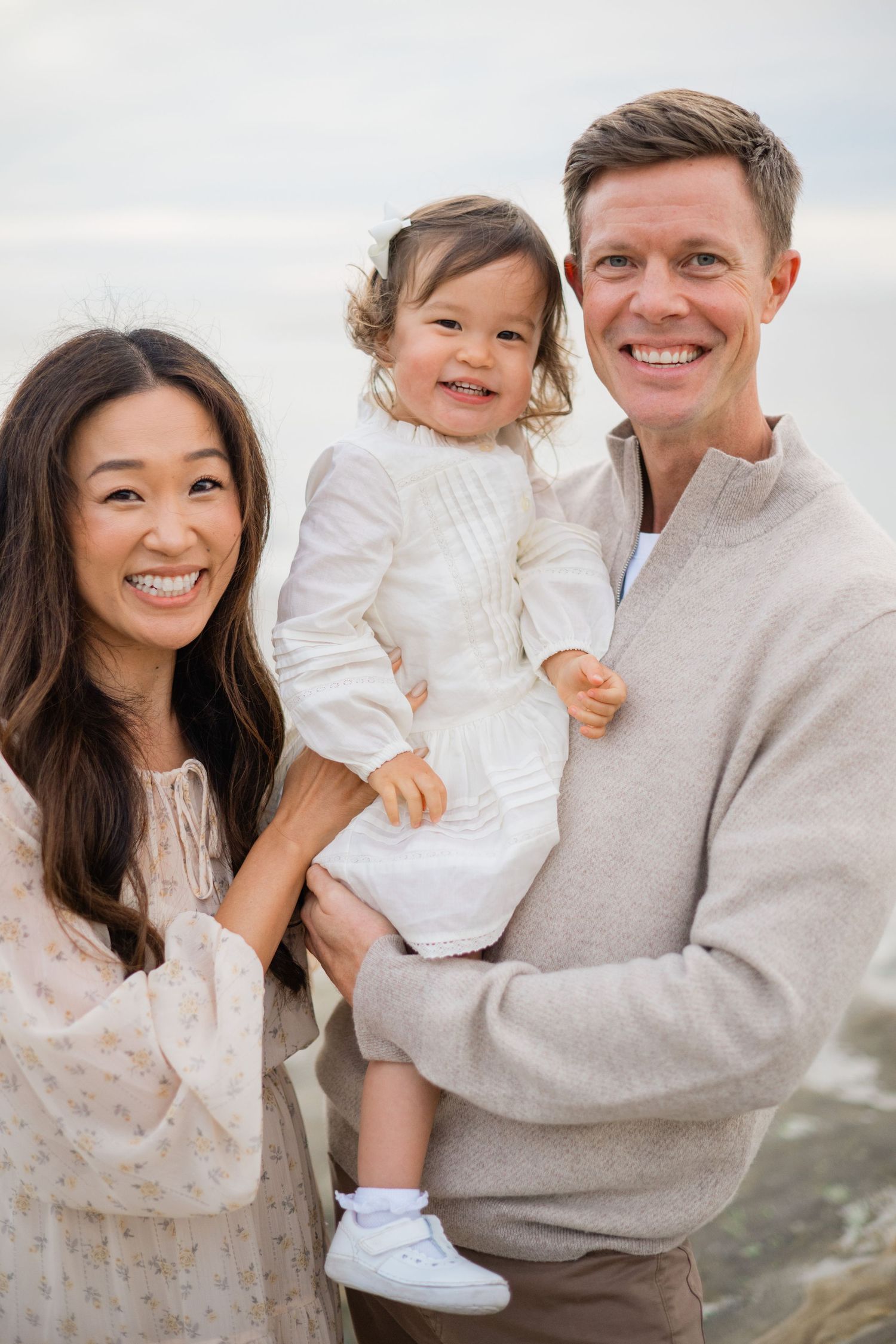 Happy family of three posing on a beach, parents smiling with their toddler daughter in a white dress.
