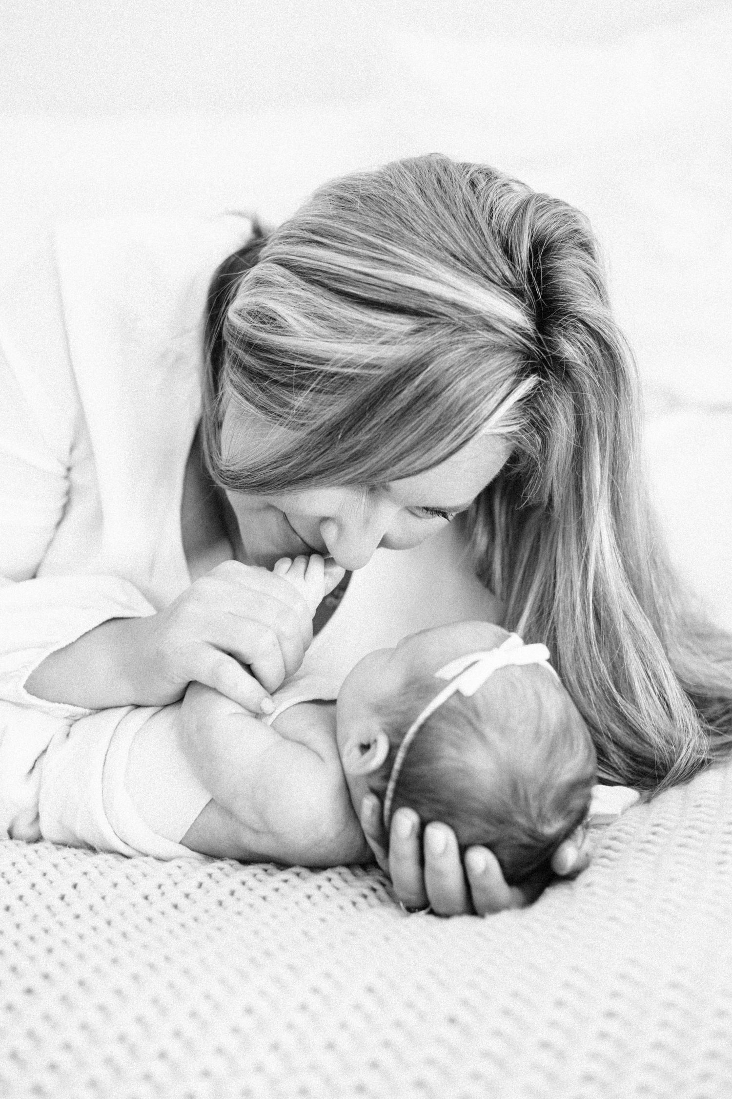 Black and white photo of a mother tenderly kissing her newborn baby while lying on a soft white bed.