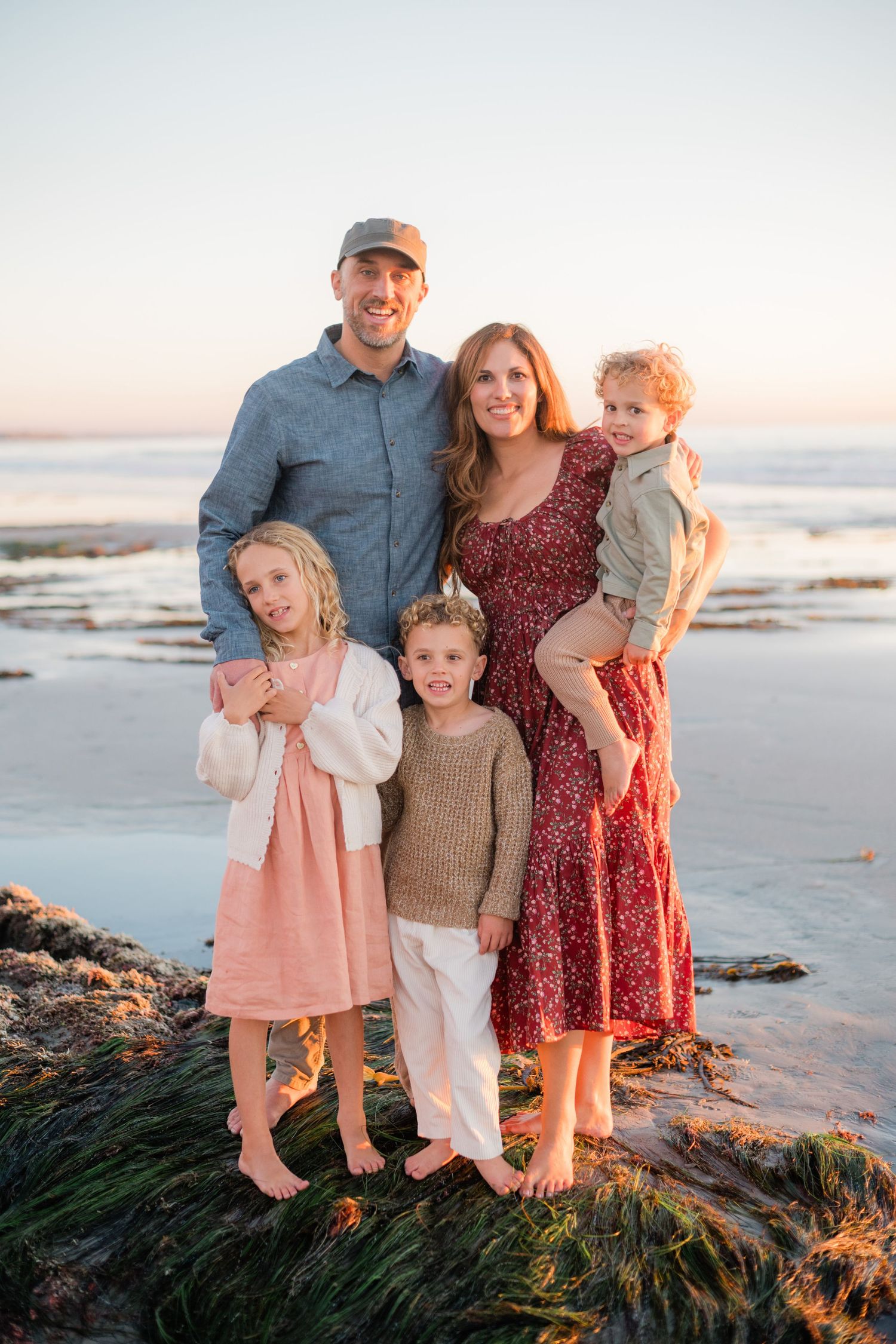 Family of five poses barefoot on rocky beach at sunset, parents with three young children in coordinated outfits.