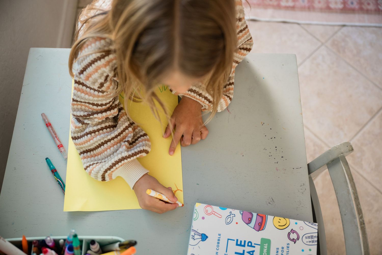 Young girl drawing on yellow paper at a table with colorful markers and a activity book nearby.