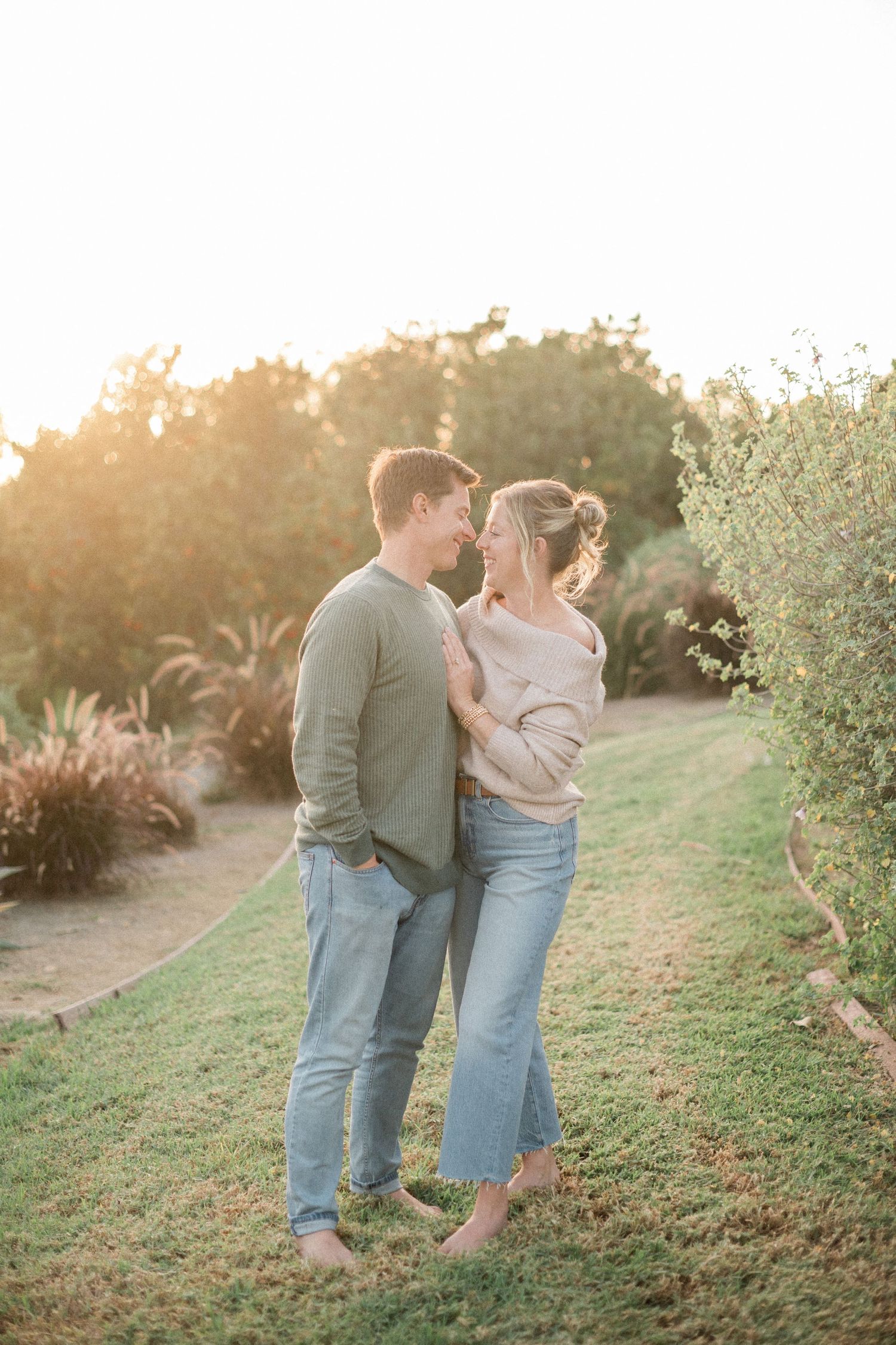 A couple embraces and smiles at each other barefoot on green grass during a golden sunset outdoors.