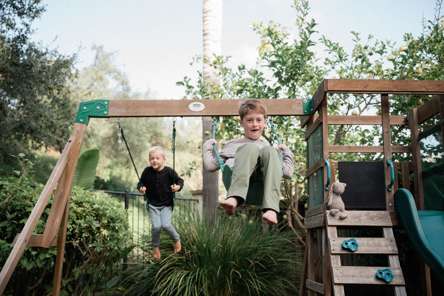 Two children playing on a wooden backyard playset with swings surrounded by lush green trees and plants.