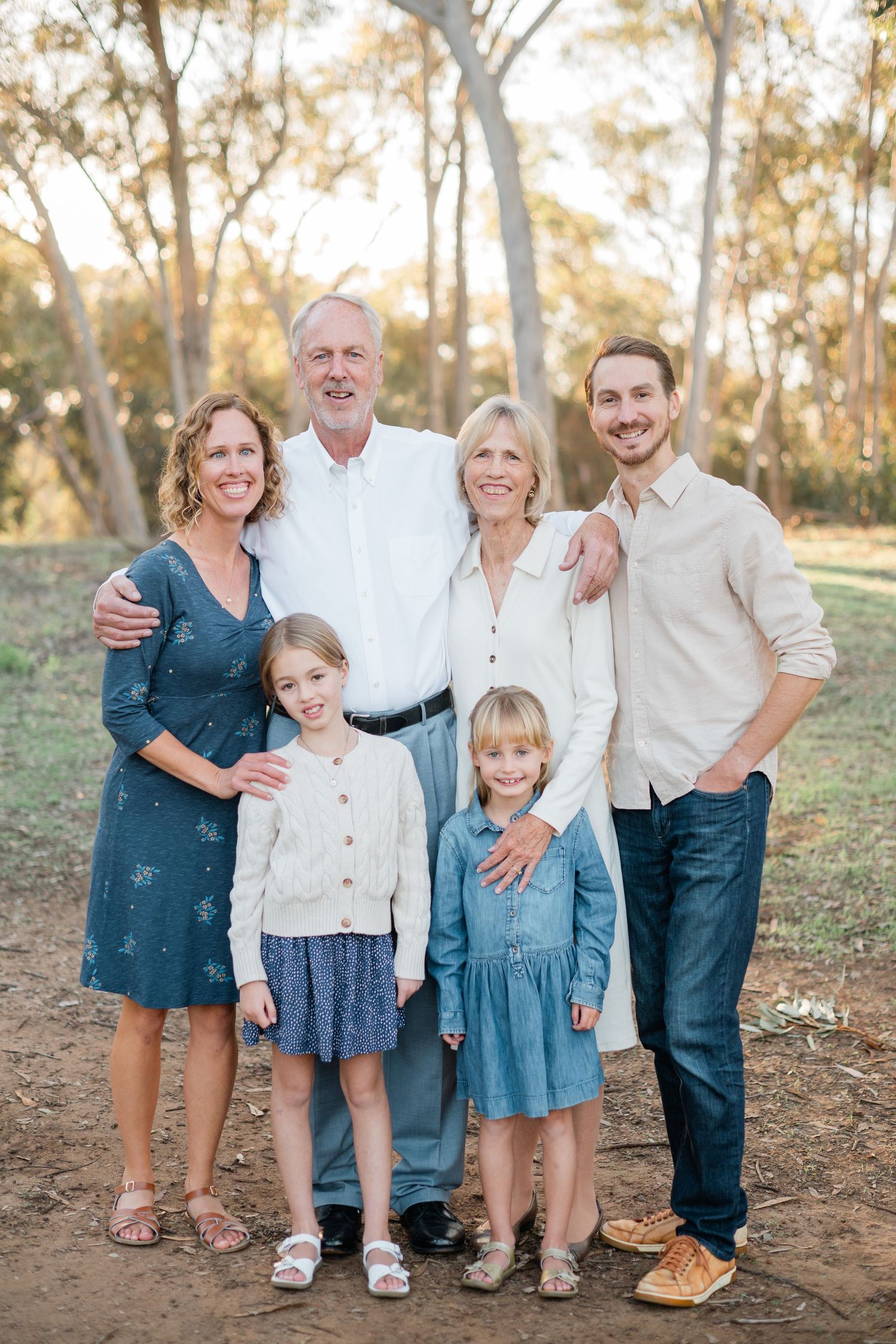 Multi-generational family of six smiling together outdoors among trees during golden hour in a warm, natural setting.