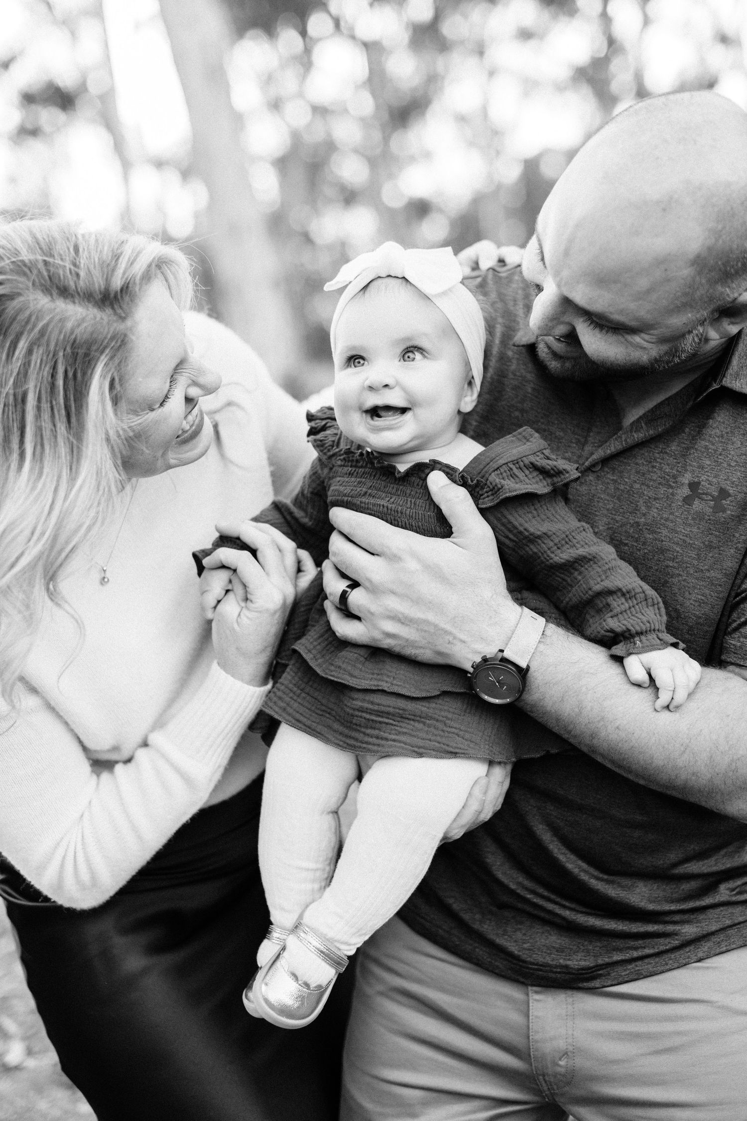 Black and white photo of parents lovingly holding their smiling baby girl outdoors among trees.