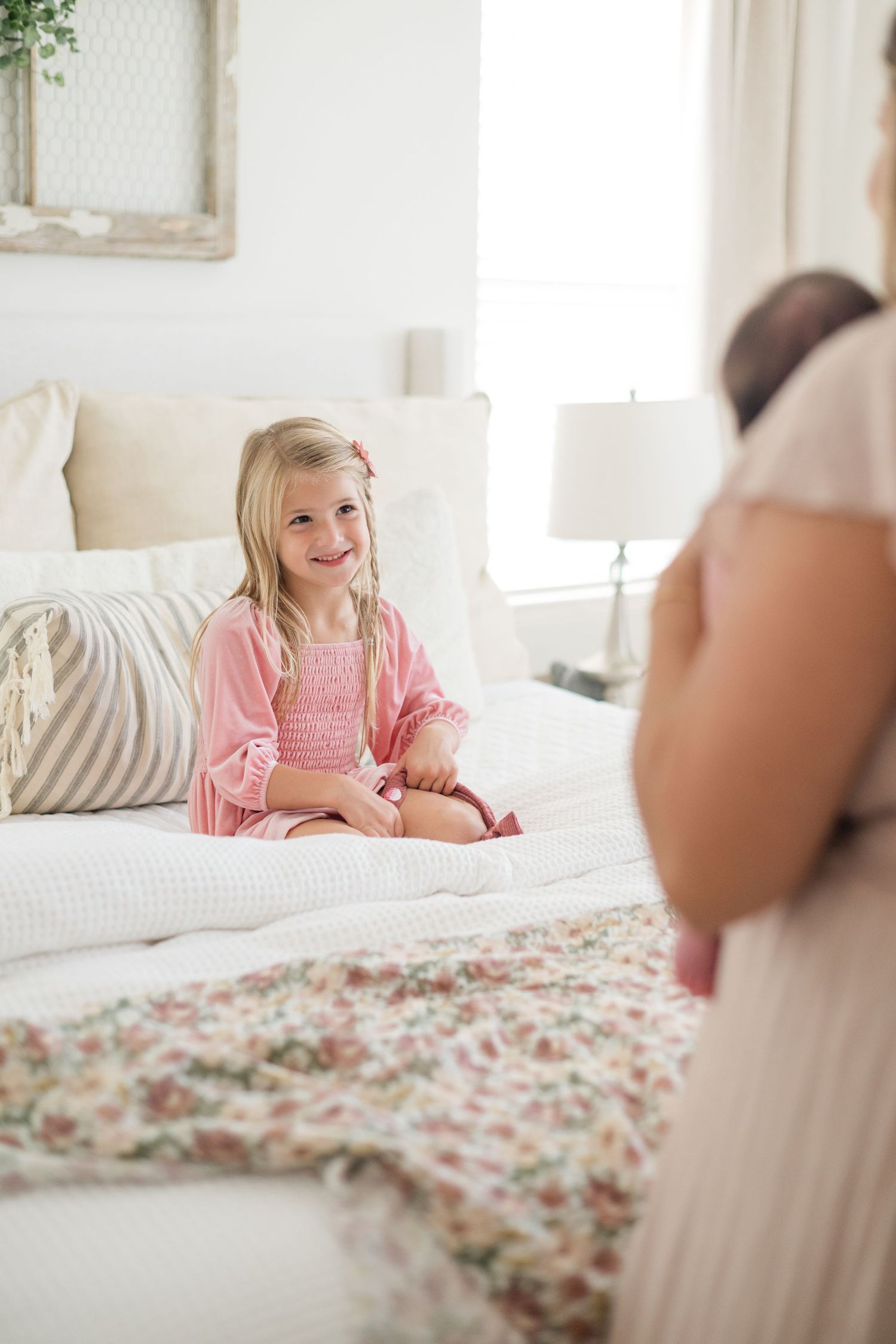 A smiling blonde girl in a pink robe sits on a white bed with floral quilt, looking at a woman holding a newborn baby.