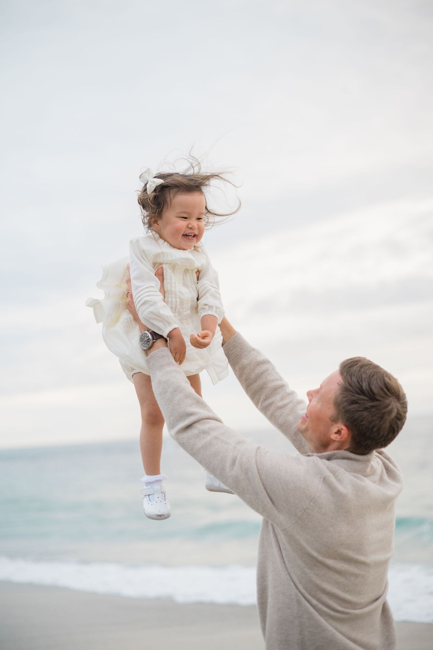 Father joyfully lifts laughing toddler girl in white dress high in the air on a beach.