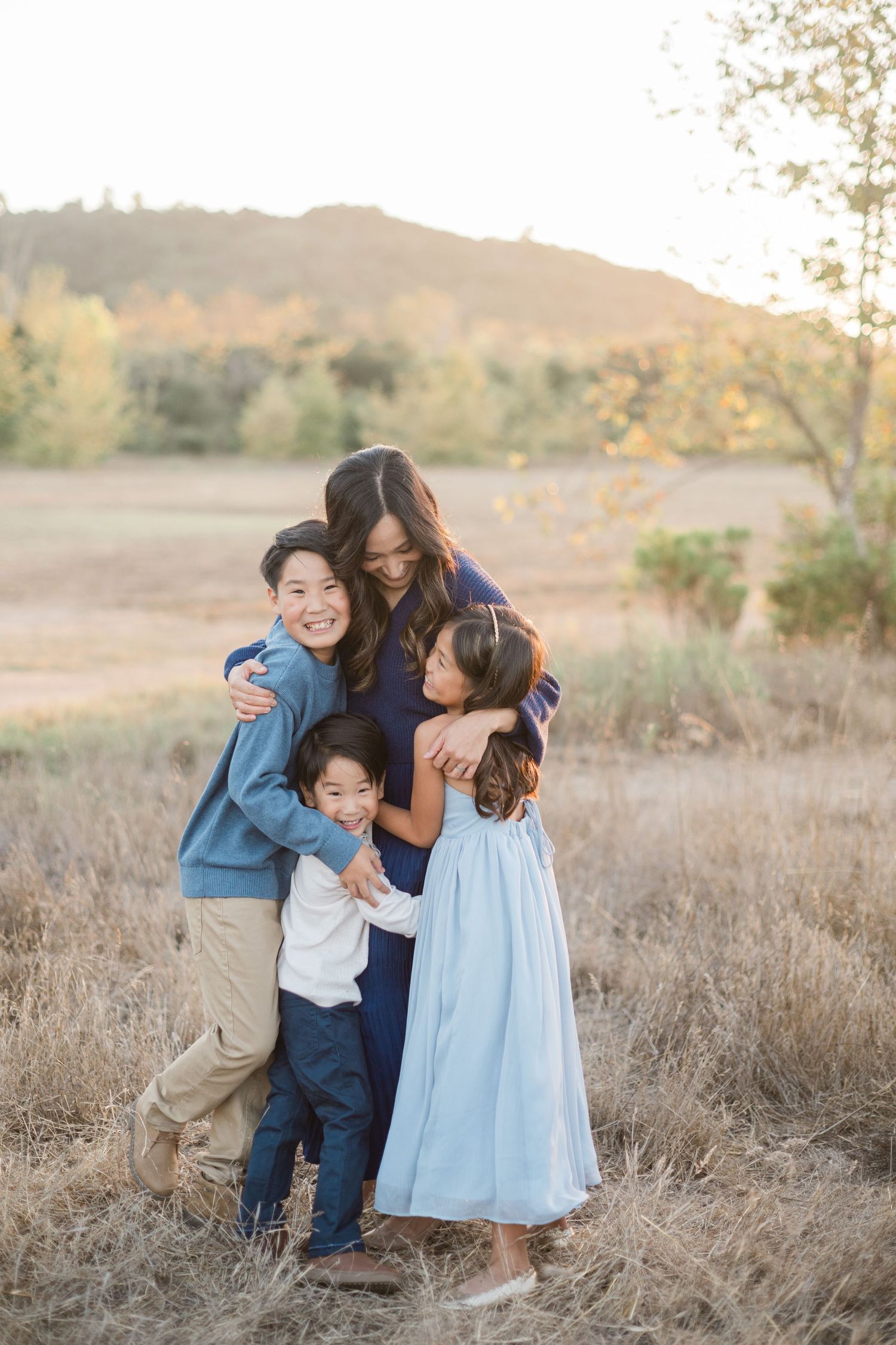 Asian mother hugging three children in sunny autumn field with colorful foliage and hills.