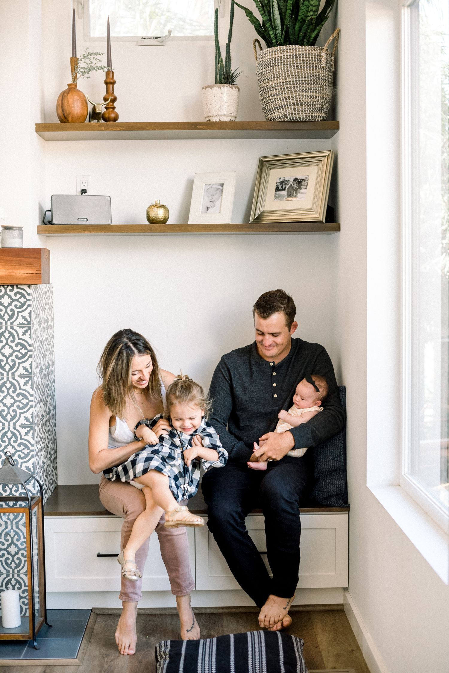 A happy family of four sits together on a bench, with parents holding their toddler and newborn baby at home.