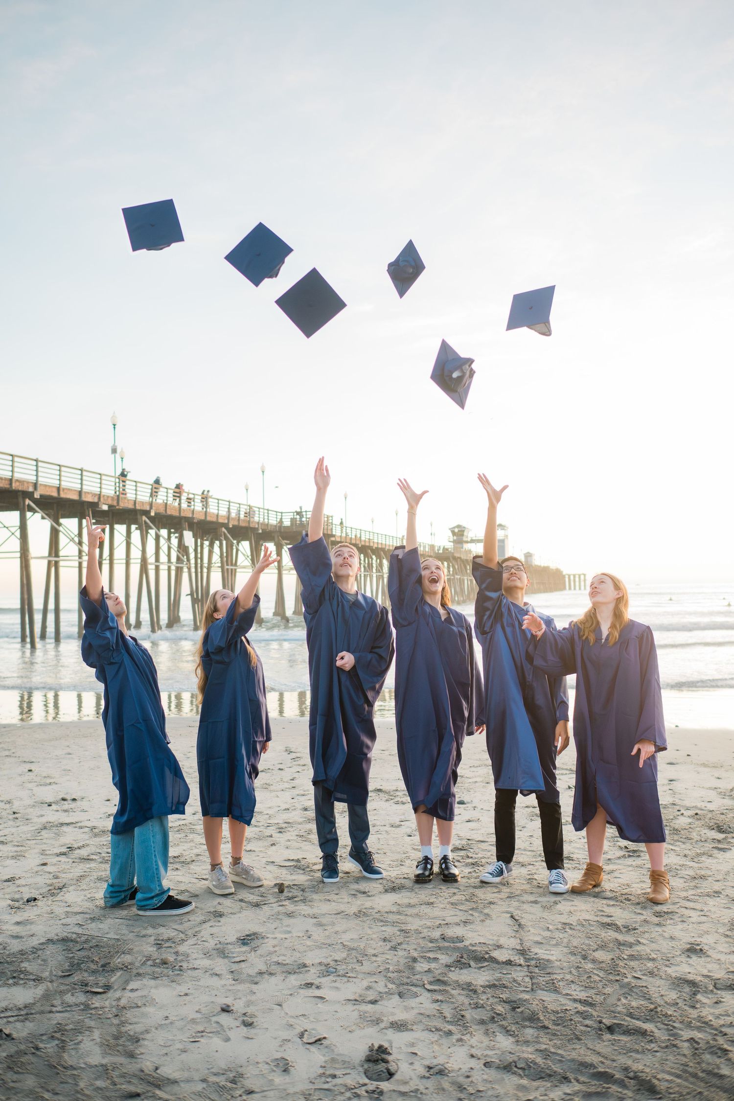 Graduates in blue gowns toss caps on a sunny beach near a pier, celebrating their academic achievement.