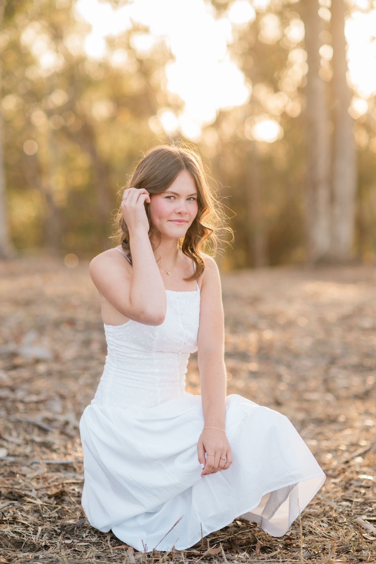 Young woman in white dress sitting outdoors in golden sunlight among trees, posing gracefully for a portrait photo.