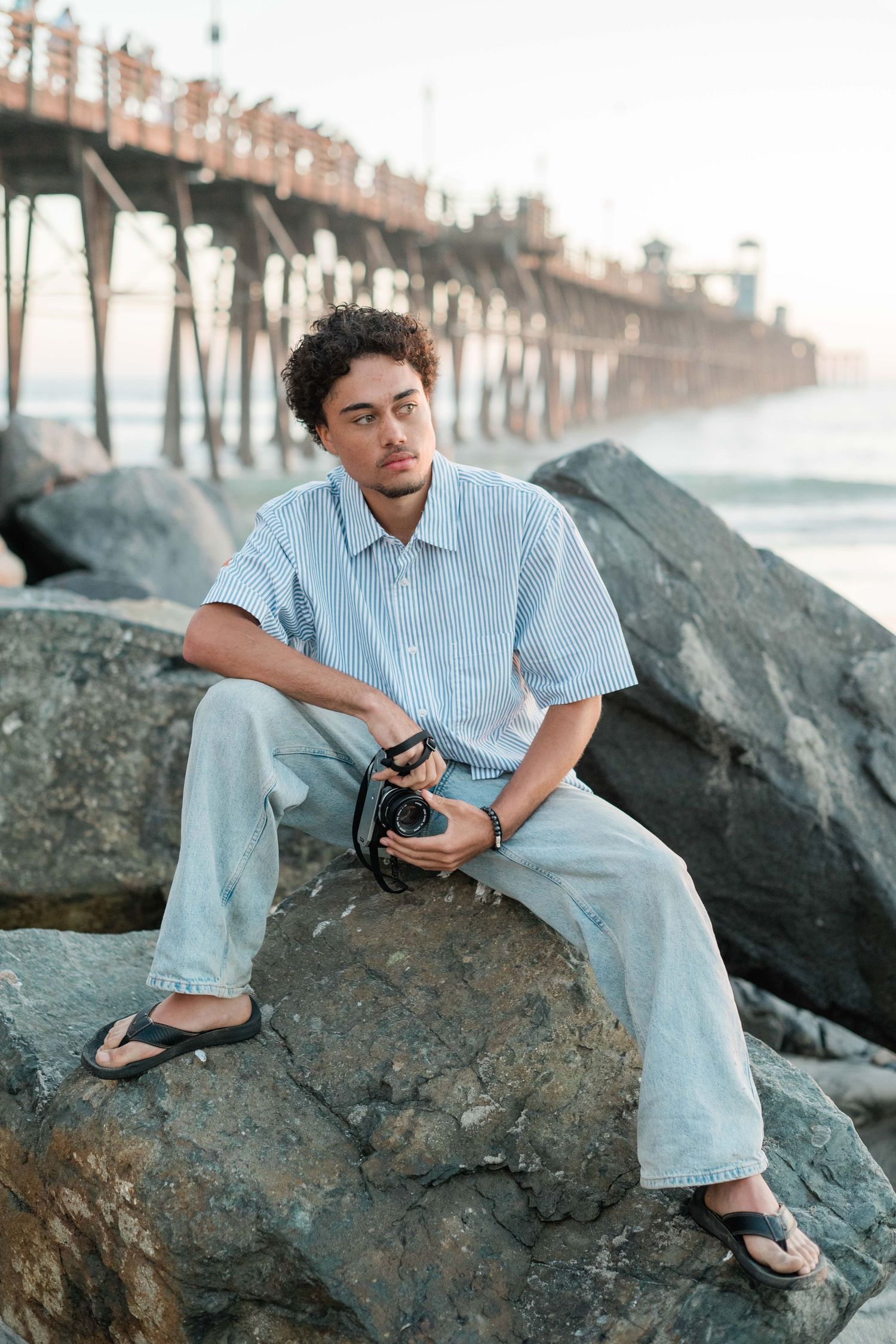 Young man with camera sitting on rocks near a beach pier, wearing a striped shirt and jeans.