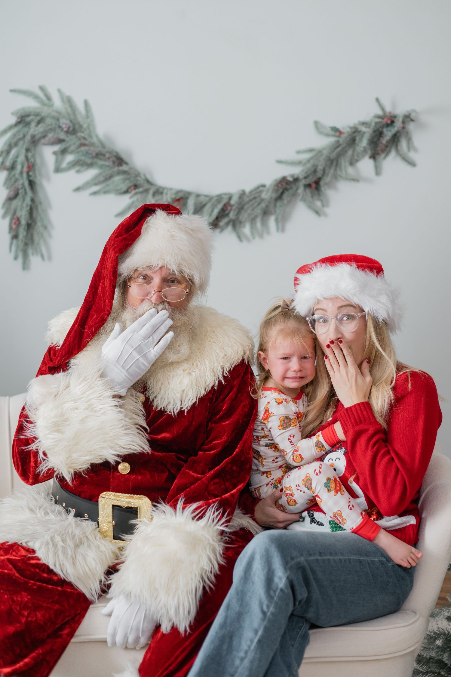 Santa and a woman in a Santa hat look shocked as a crying toddler sits between them on a couch.