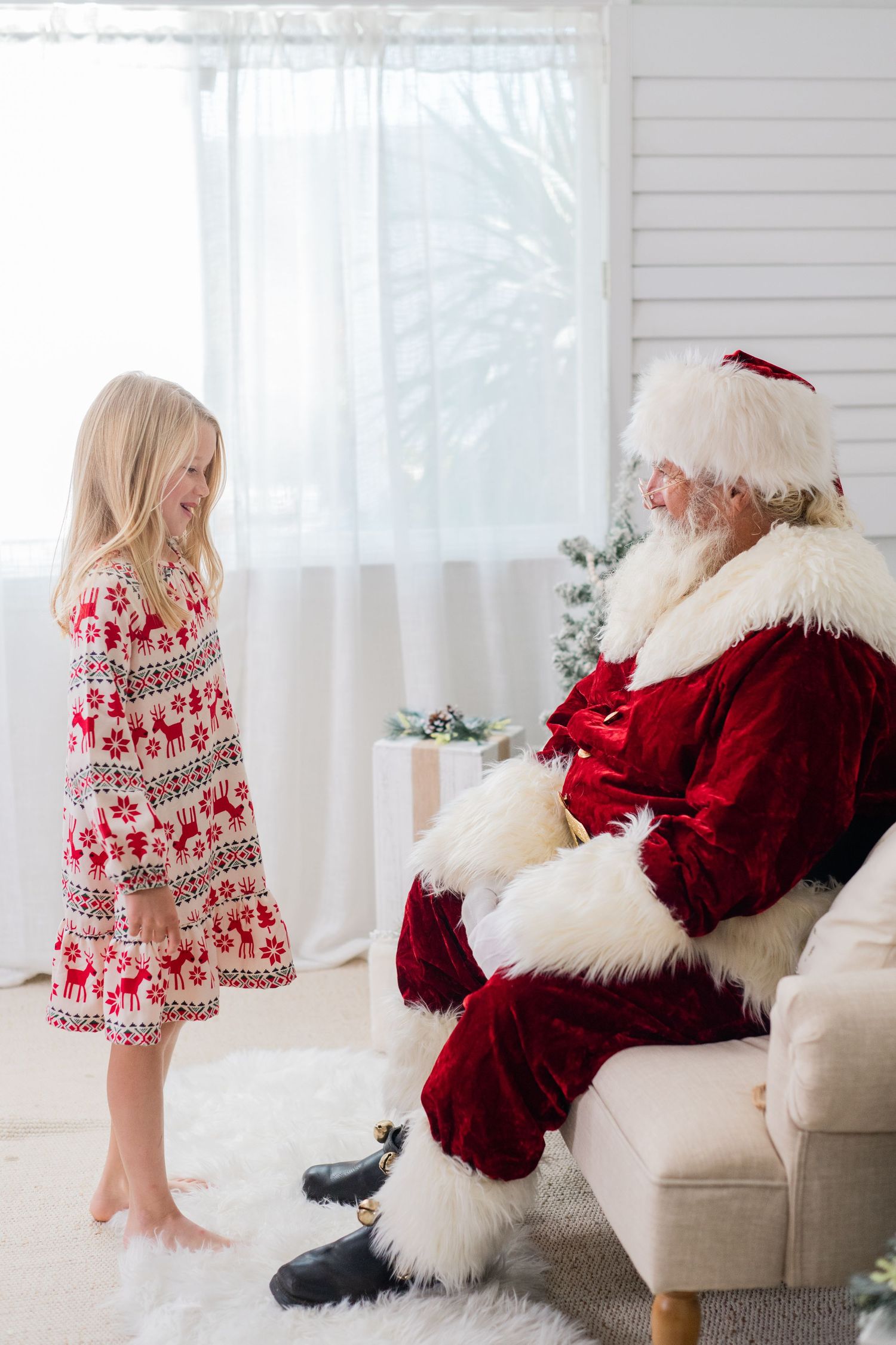 A young girl in a reindeer print dress smiles at Santa Claus seated in a chair during a Christmas photo session.