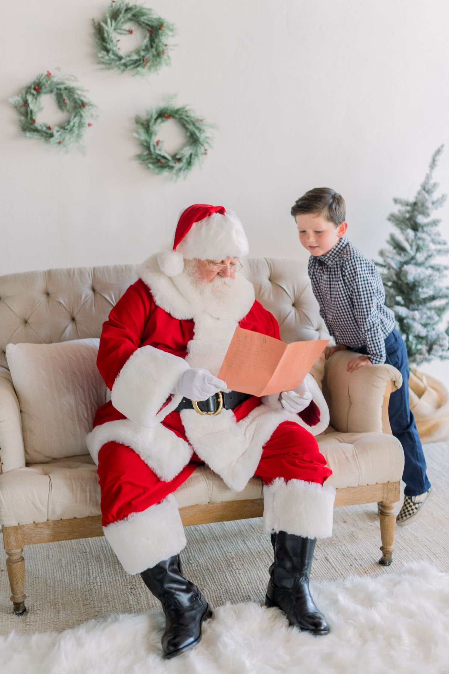 Santa Claus reads a letter on a cozy sofa while a young boy in plaid shirt watches during Christmas photos.
