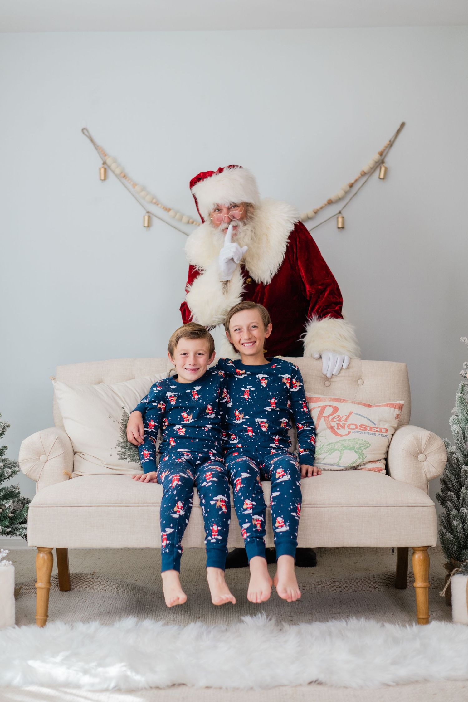 Two boys in matching Santa pajamas sit on a cream sofa with Santa Claus behind them in a festive Christmas setting.
