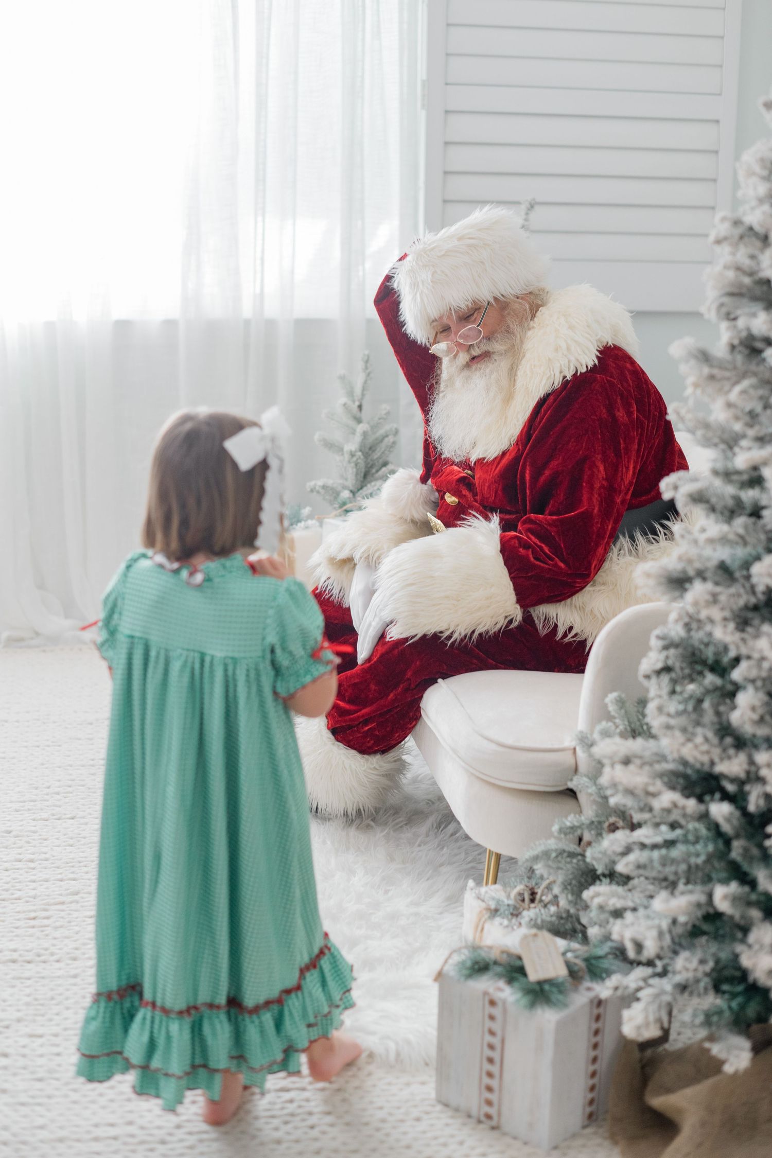 A little girl in a teal dress listens to Santa Claus beside a frosted Christmas tree indoors.