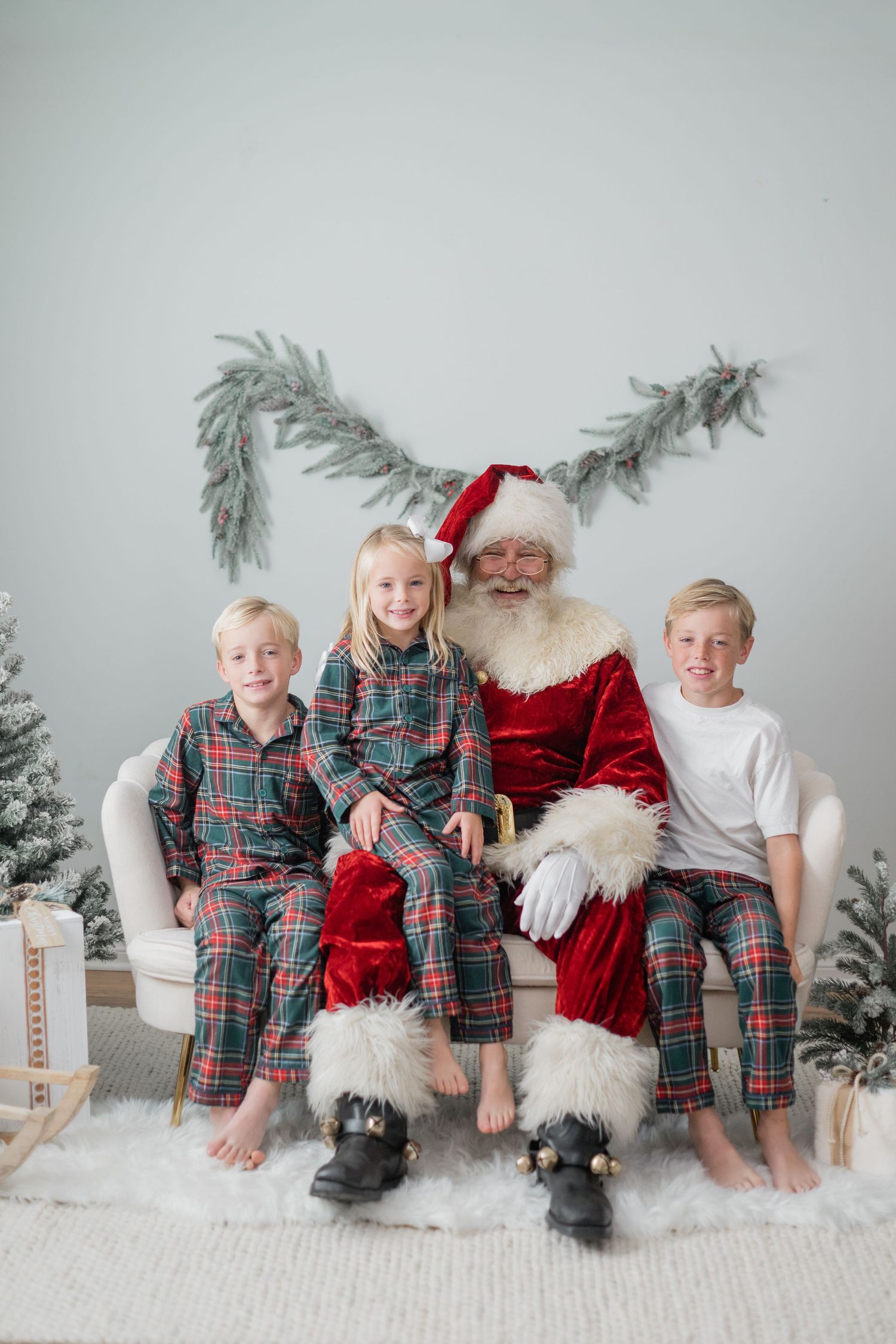 Santa Claus sits on a white chair with three children in plaid pajamas, surrounded by Christmas trees and festive decor.