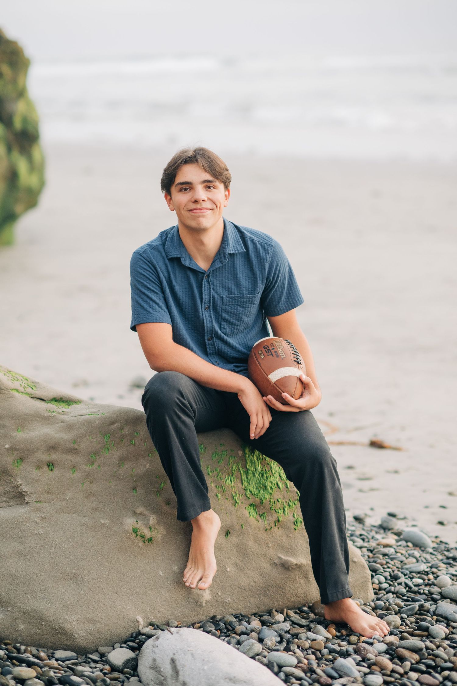 Young man in blue shirt sitting barefoot on a rock holding a football on a pebble beach.