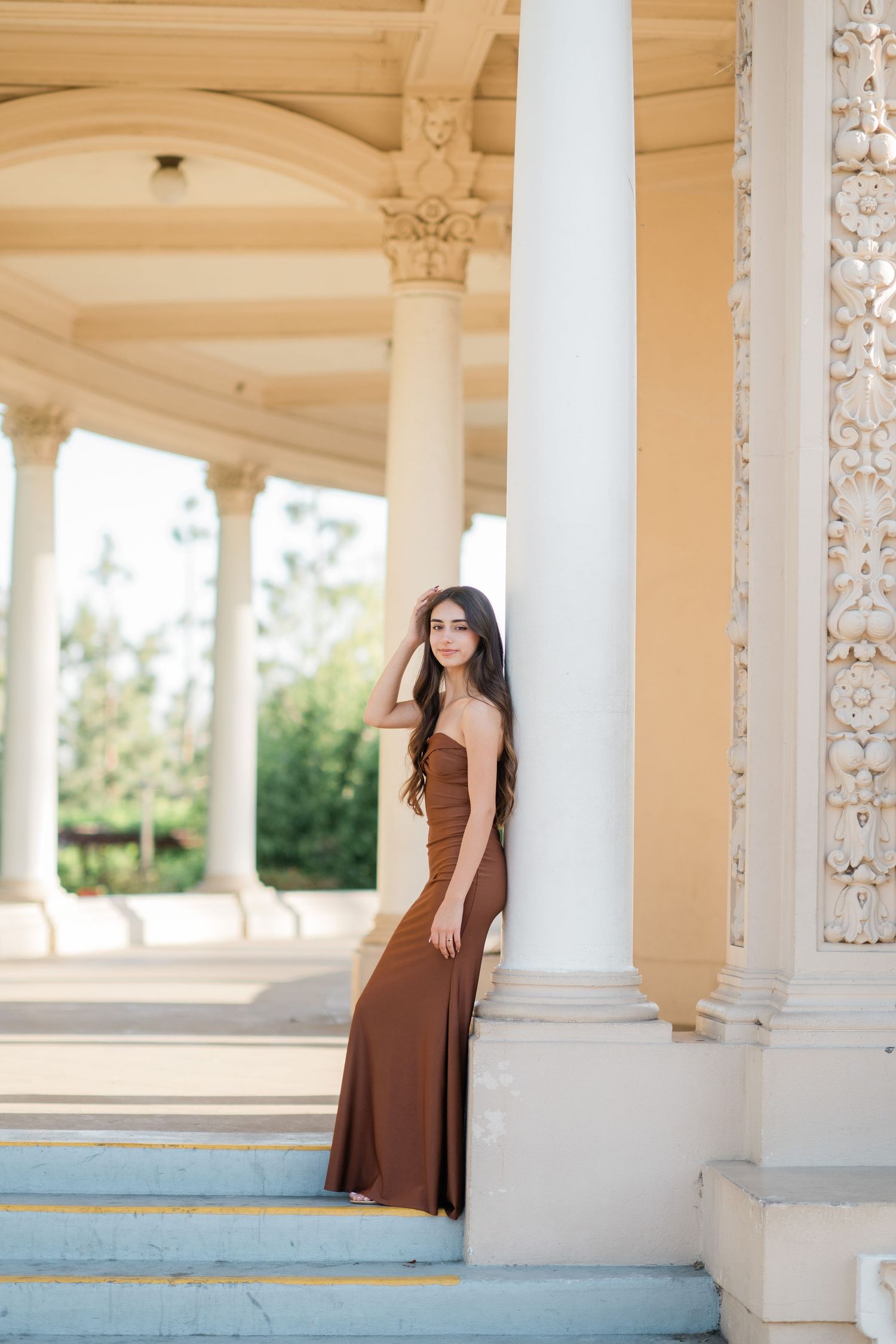 Young woman in brown strapless gown poses gracefully between white columns of an elegant classical architecture.