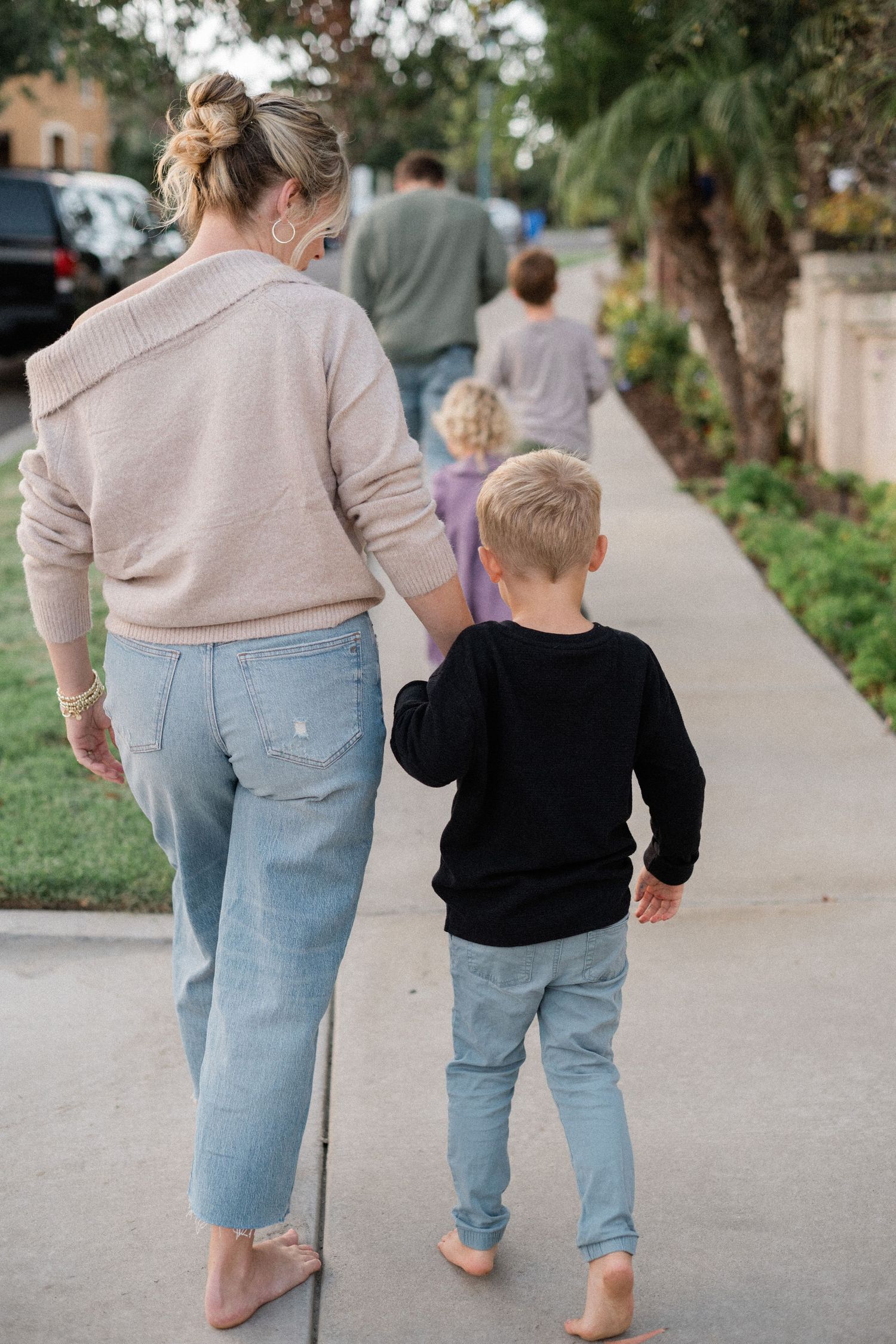 Mother and young boy in black sweatshirt walking barefoot on a suburban sidewalk, holding hands from behind.