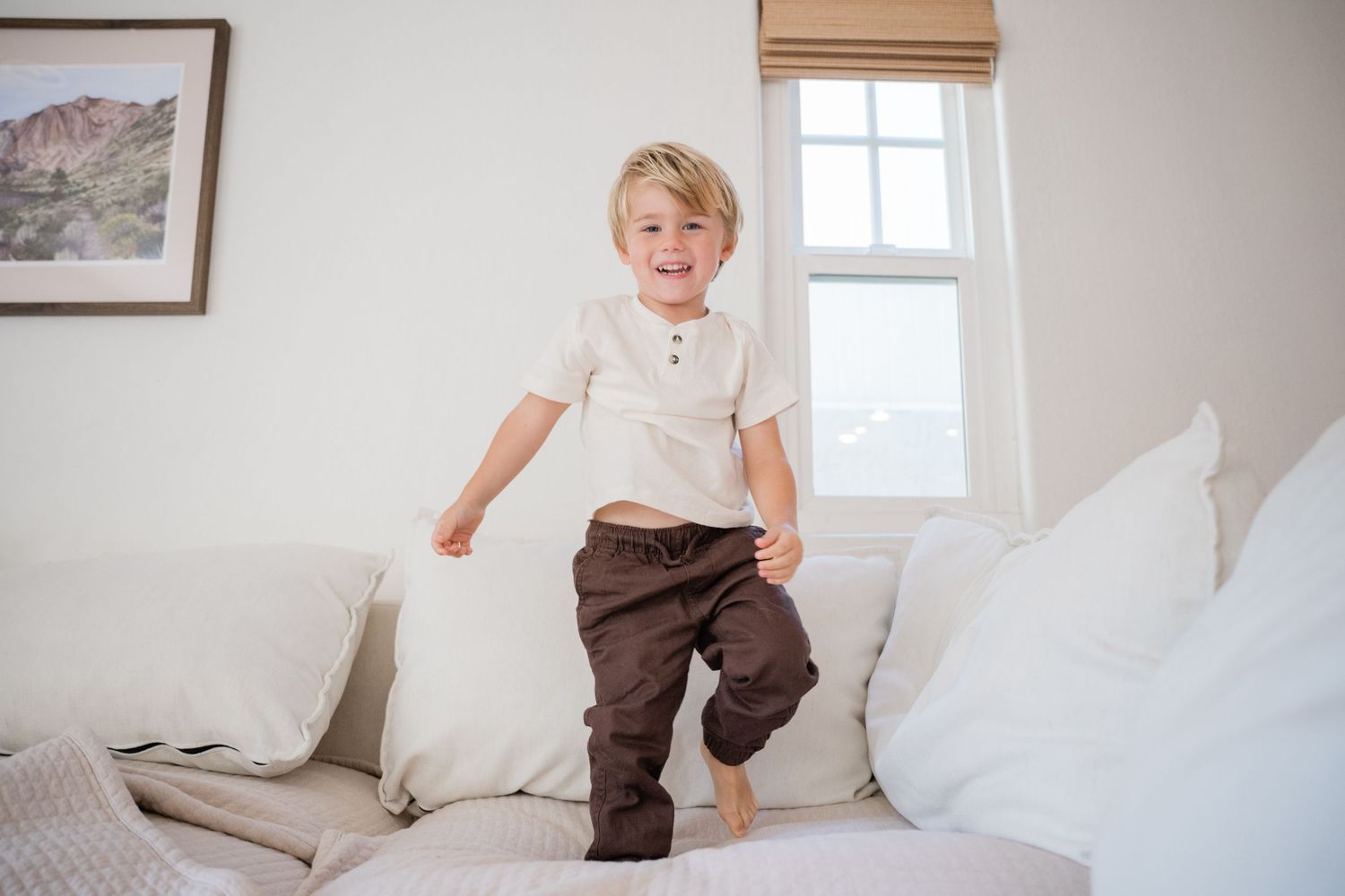 Young boy jumping on white sofa cushions in bright living room, wearing cream shirt and brown pants.