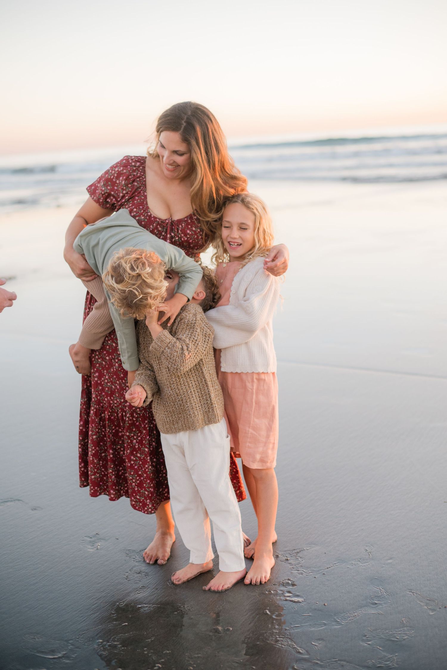 A mother embraces her three children on a sandy beach at sunset, all barefoot in warm-toned outfits.