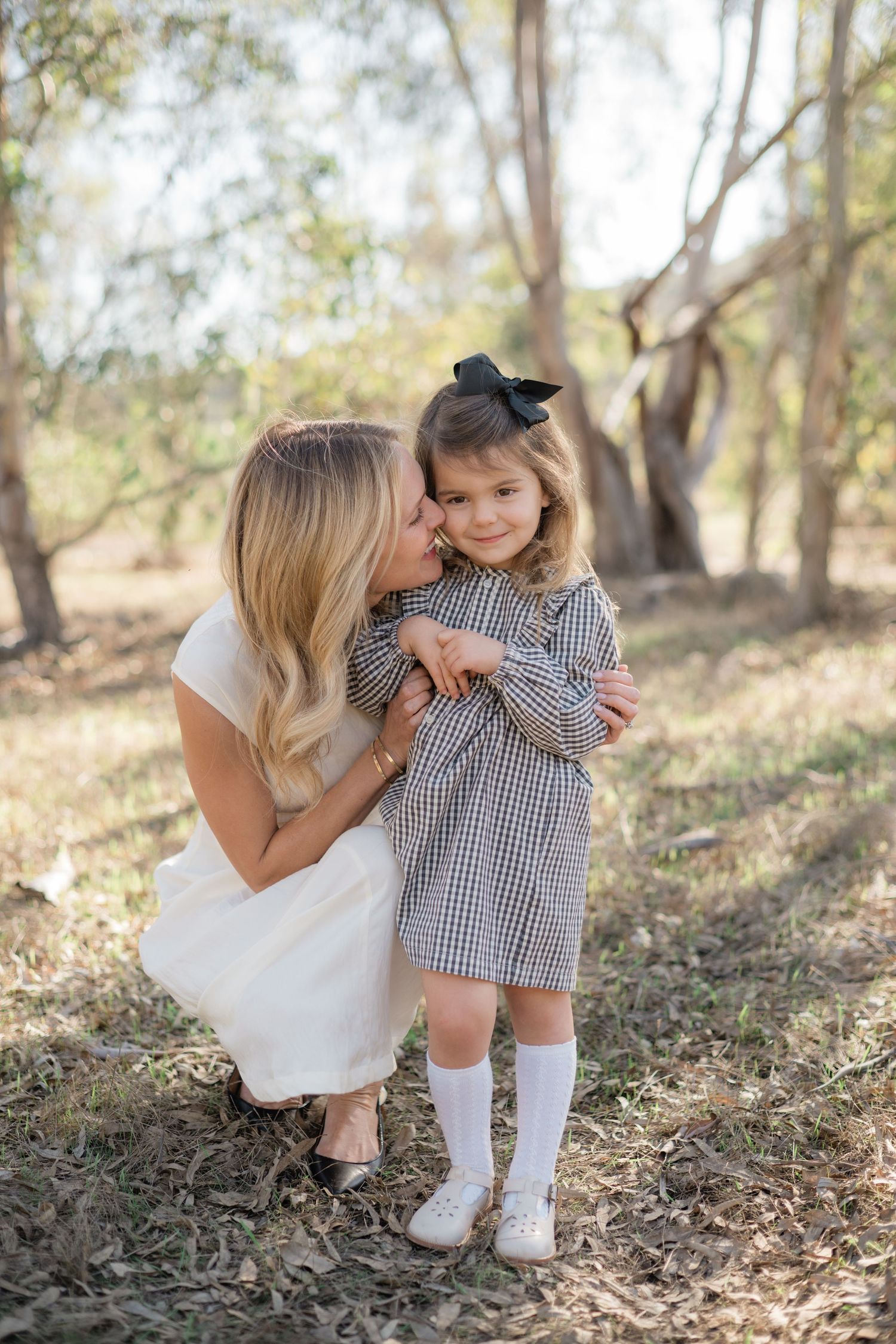 A loving mother kisses her young daughter wearing a gingham dress and black bow outdoors.