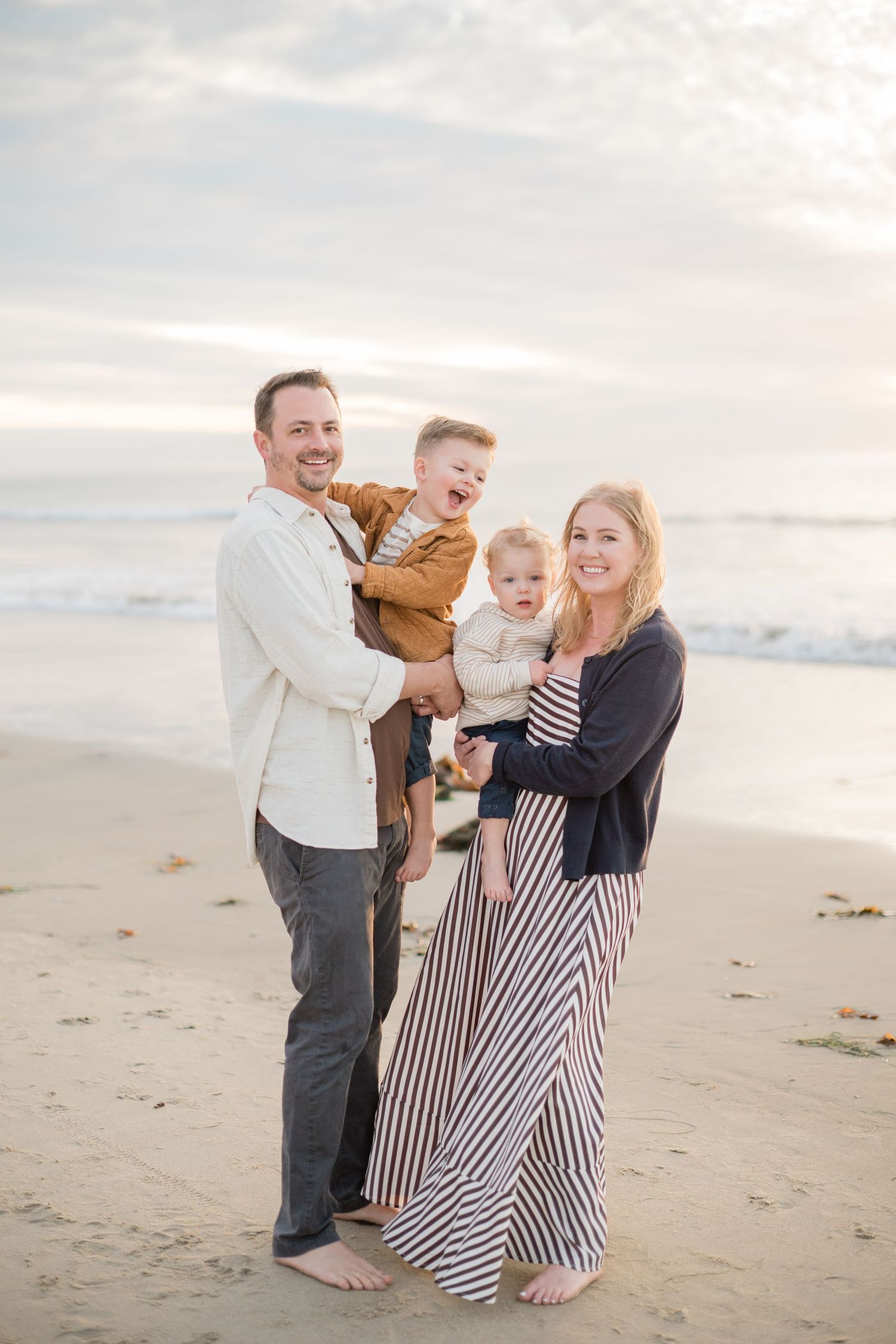 Happy family of four posing on a sandy beach at sunset, parents holding two young children while smiling.