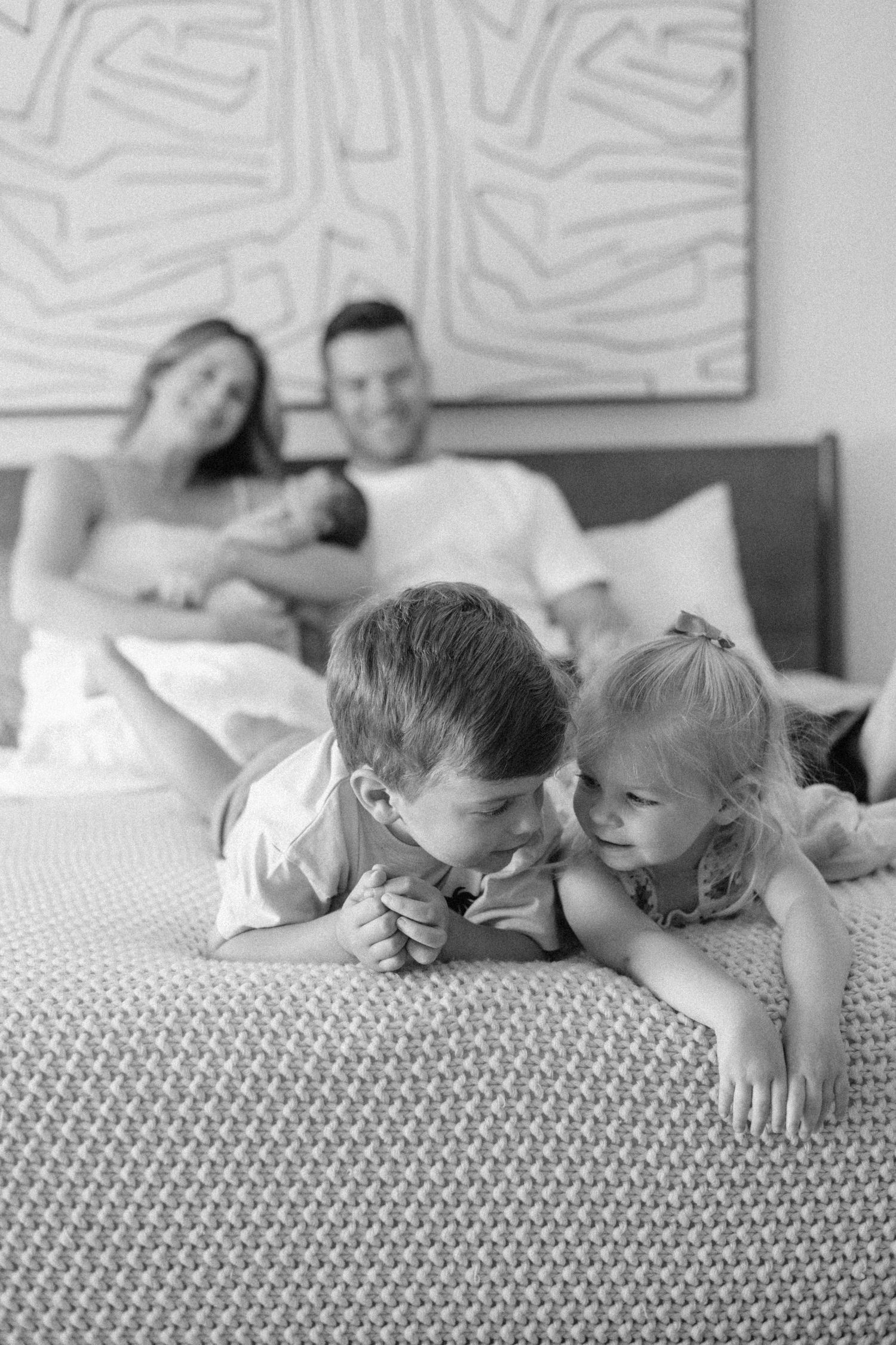 Black and white family photo with two children playing on bed while parents hold newborn baby in background.