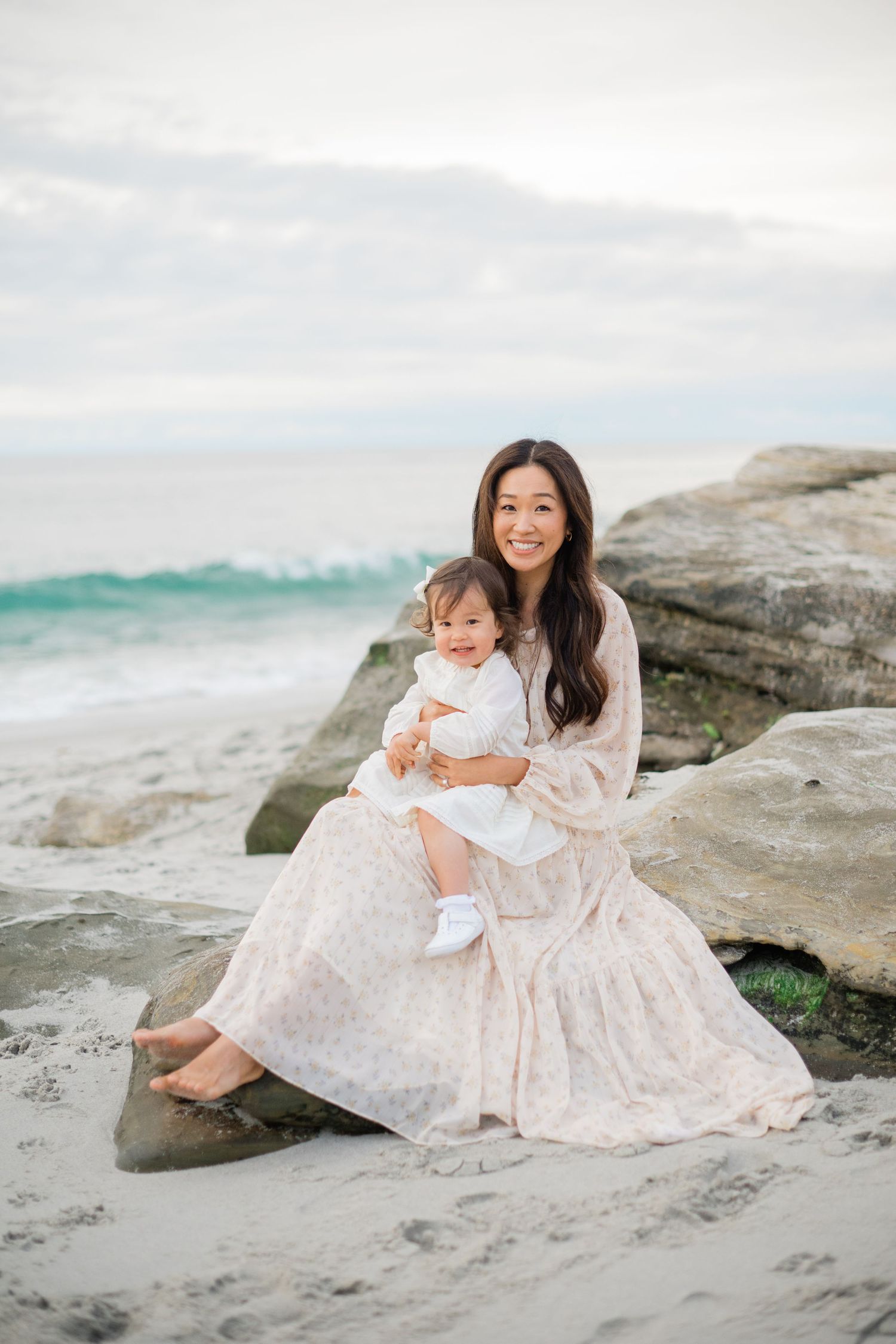 A woman in a floral dress sits on beach rocks holding a baby in white, with ocean waves in the background.