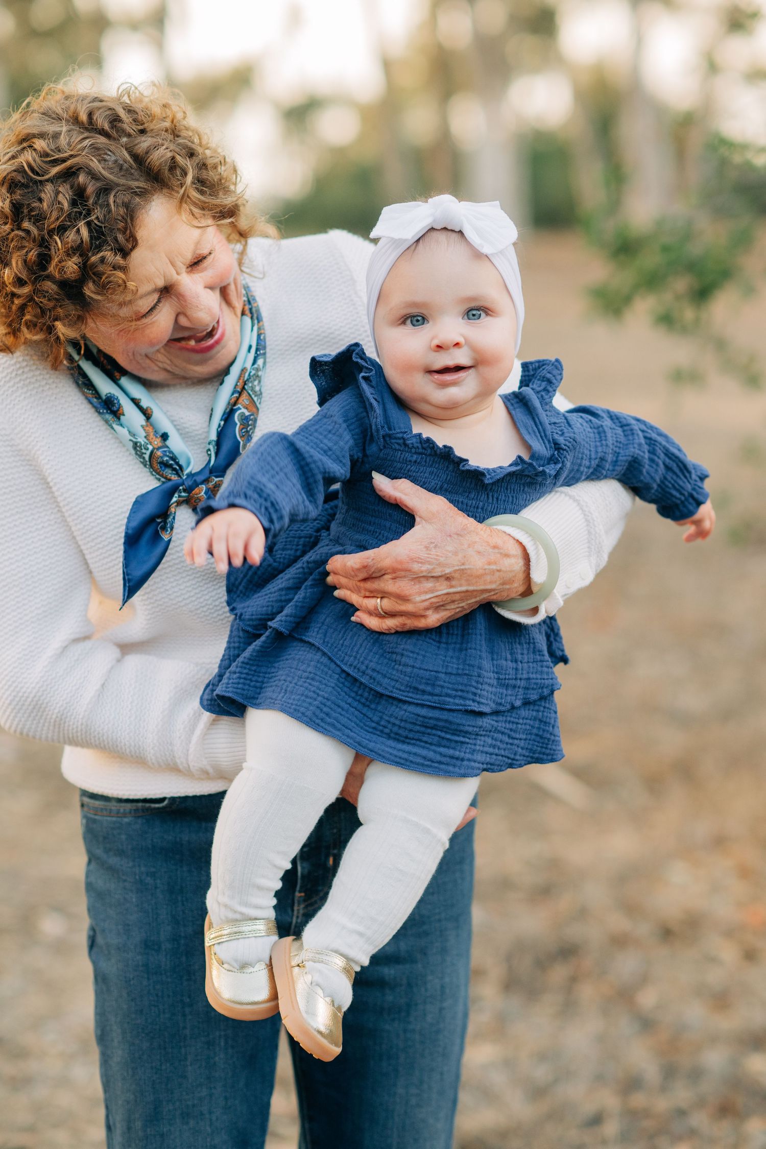 Grandmother with curly hair holds smiling blue-eyed baby in blue dress and white bow outdoors.