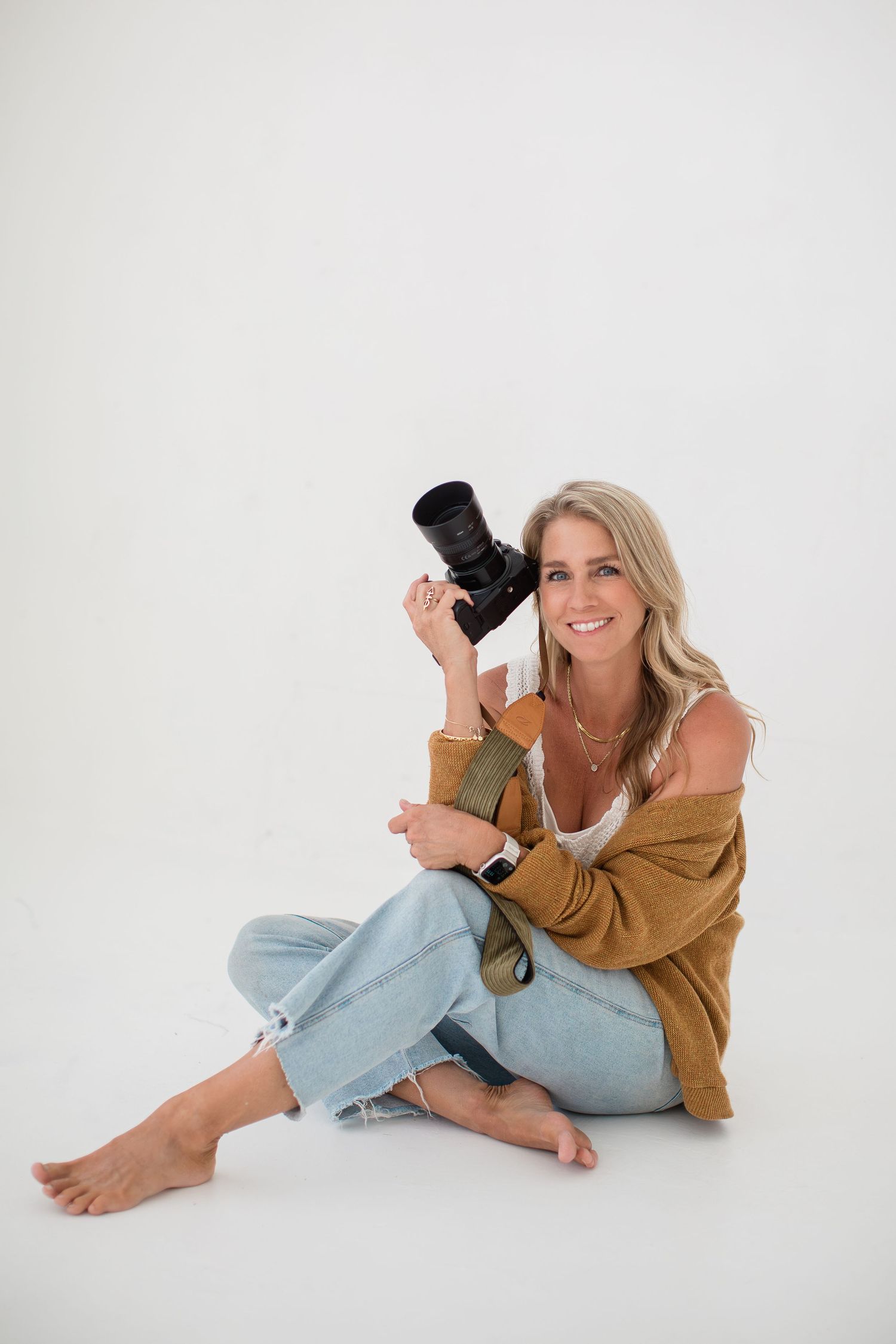 Smiling blonde woman sitting on white background holding a camera, wearing jeans and a mustard cardigan.
