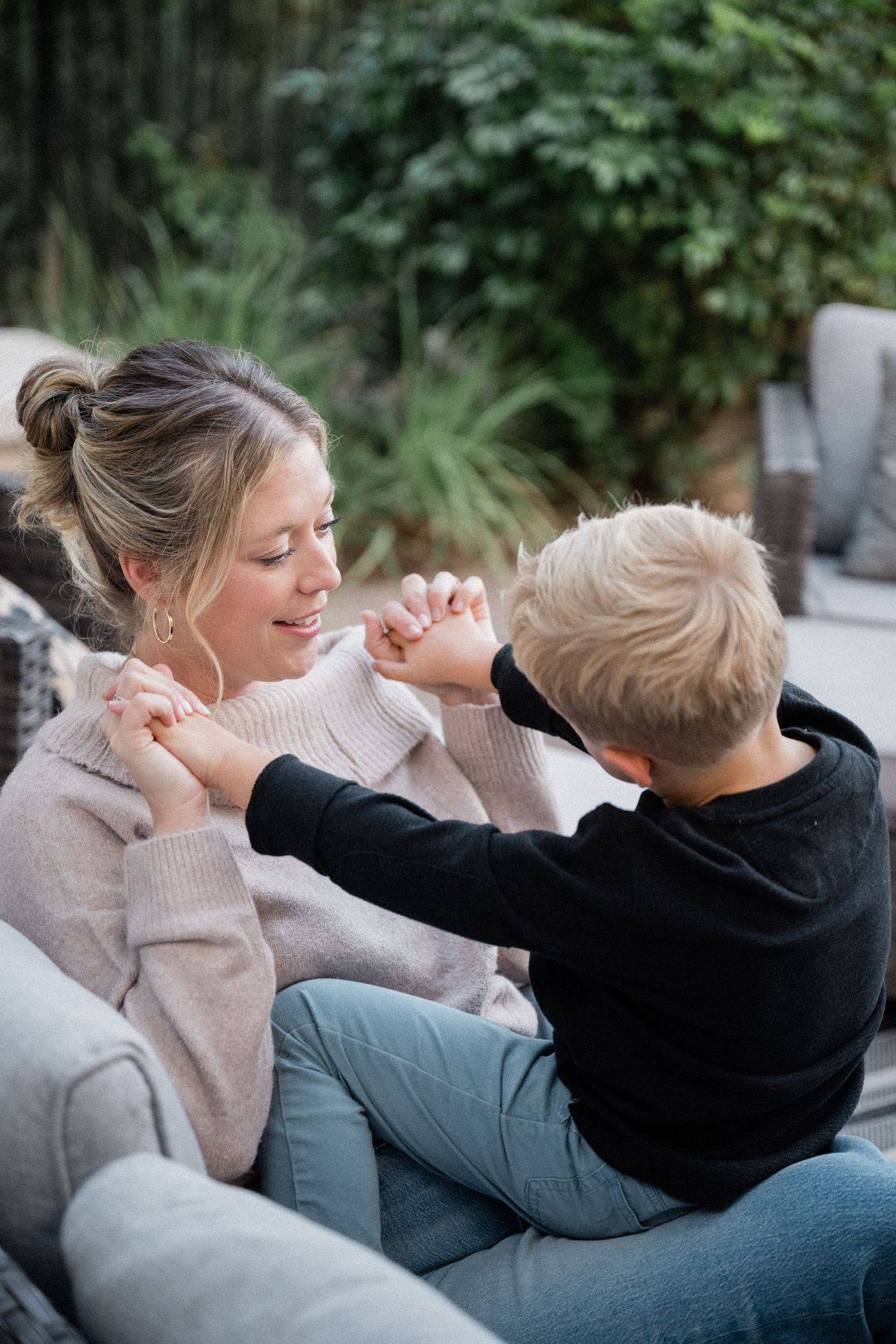 A smiling blonde mother in a beige sweater playfully interacts with her young son on an outdoor patio sofa.