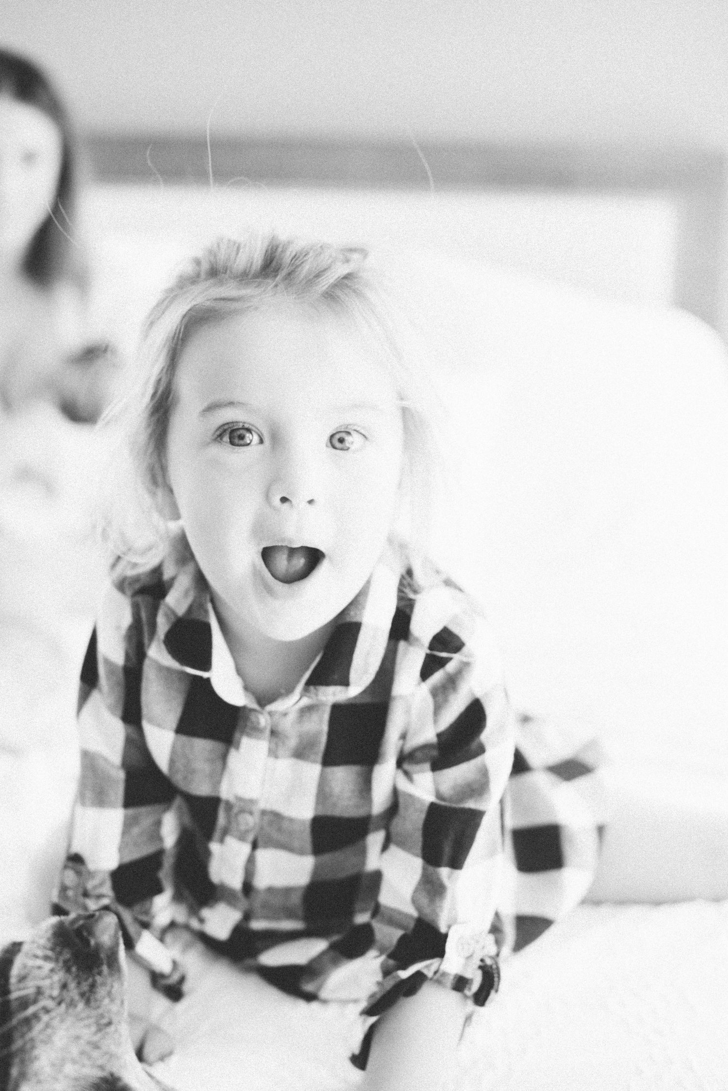 Adorable toddler girl in plaid shirt making a surprised face while sitting on a white bed.