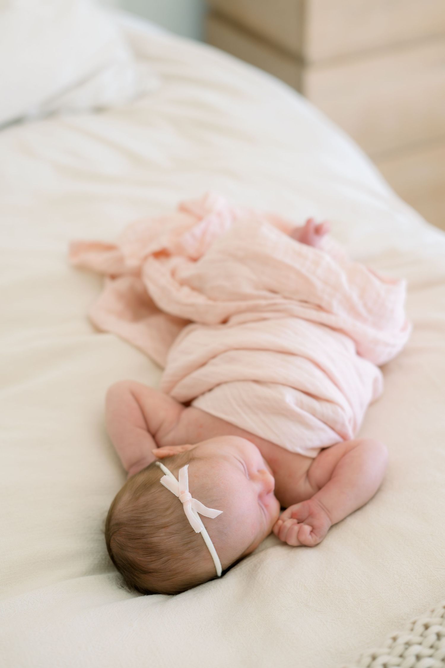 Newborn baby sleeping on white bed wrapped in pink muslin swaddle blanket with white bow headband.