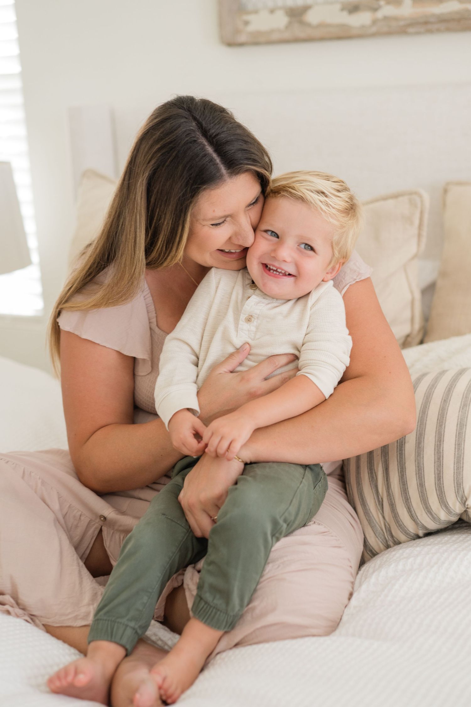 A loving mother cuddles her smiling toddler boy on a bed in a bright, cozy bedroom.