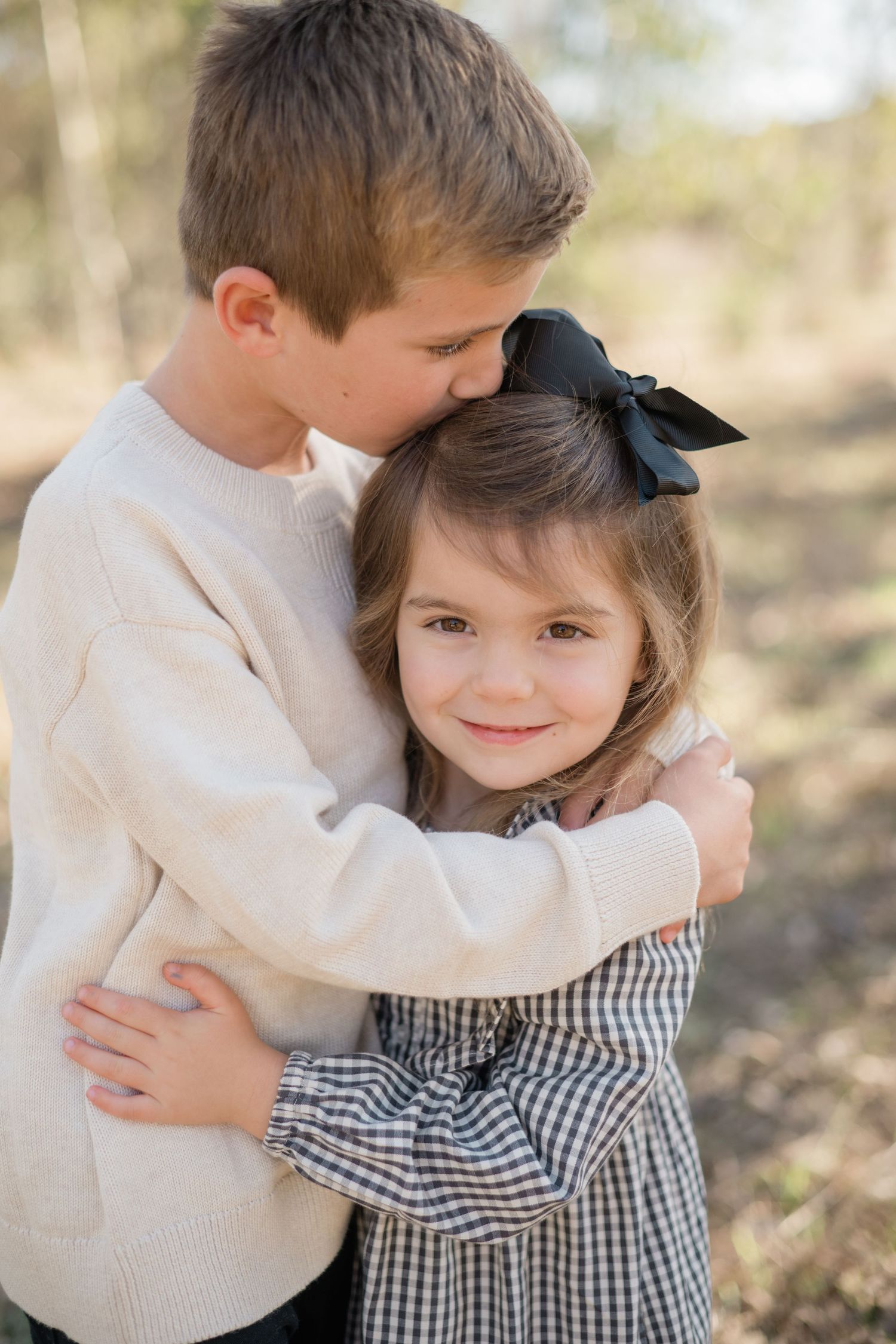 A boy in a cream sweater hugs a smiling girl in a plaid dress with a black bow outdoors.