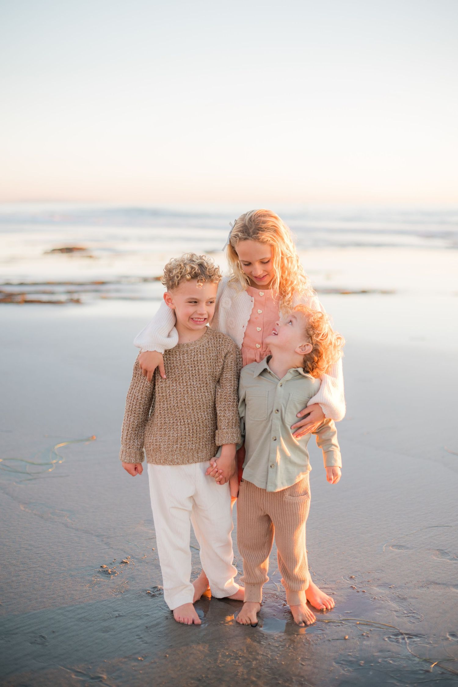 A mother and her two curly-haired children embrace on a sandy beach at sunset, barefoot and smiling.