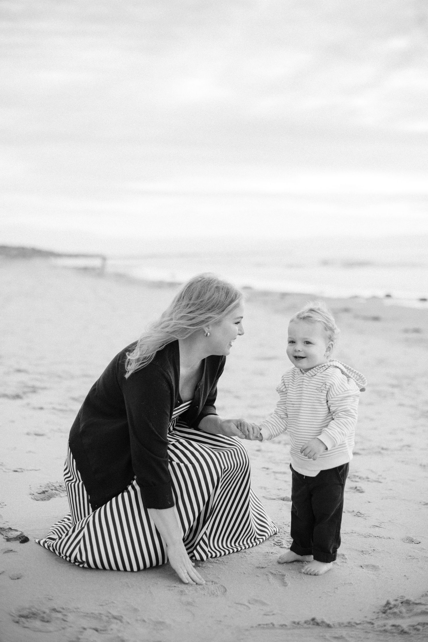 Black and white photo of a mother in a striped skirt kneeling on the beach with her toddler.