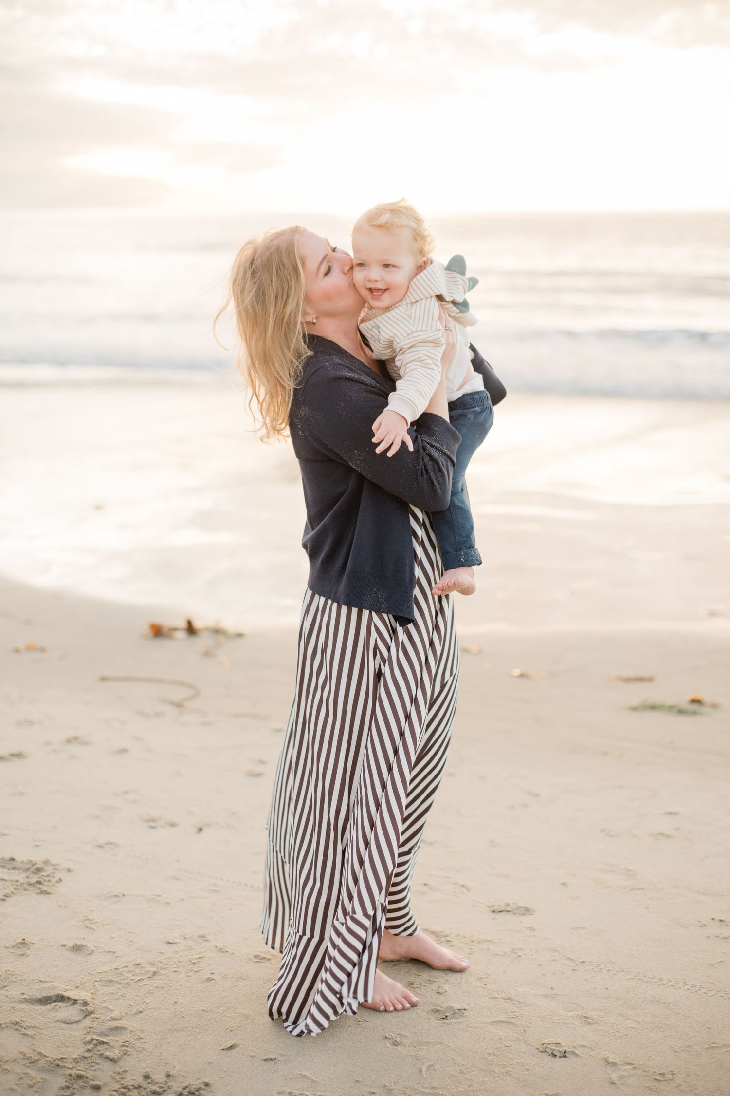 Blonde mother holding laughing baby at sunset beach, wearing striped maxi skirt.