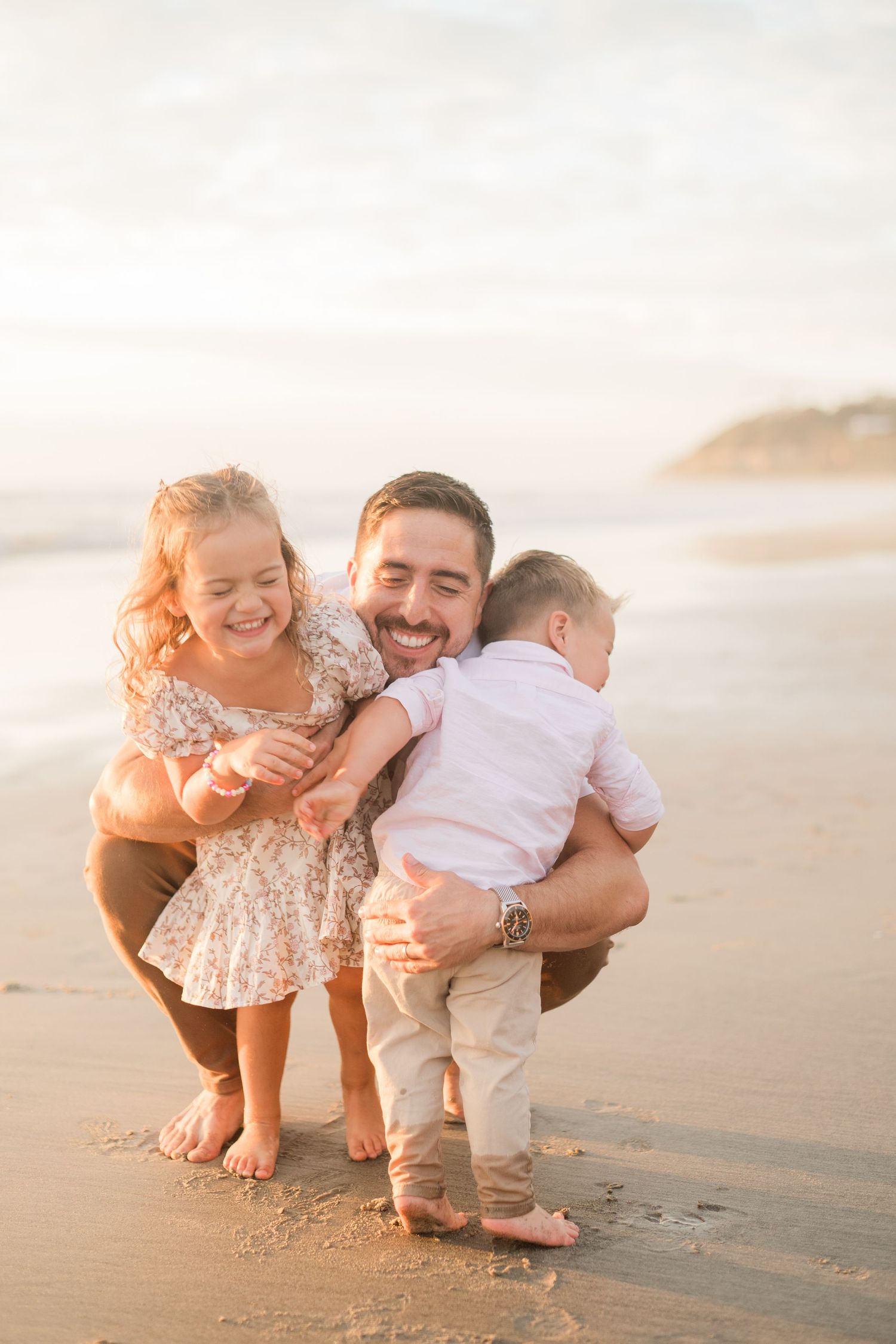 A smiling father crouches on a sandy beach, joyfully hugging his young daughter and toddler son at golden hour.