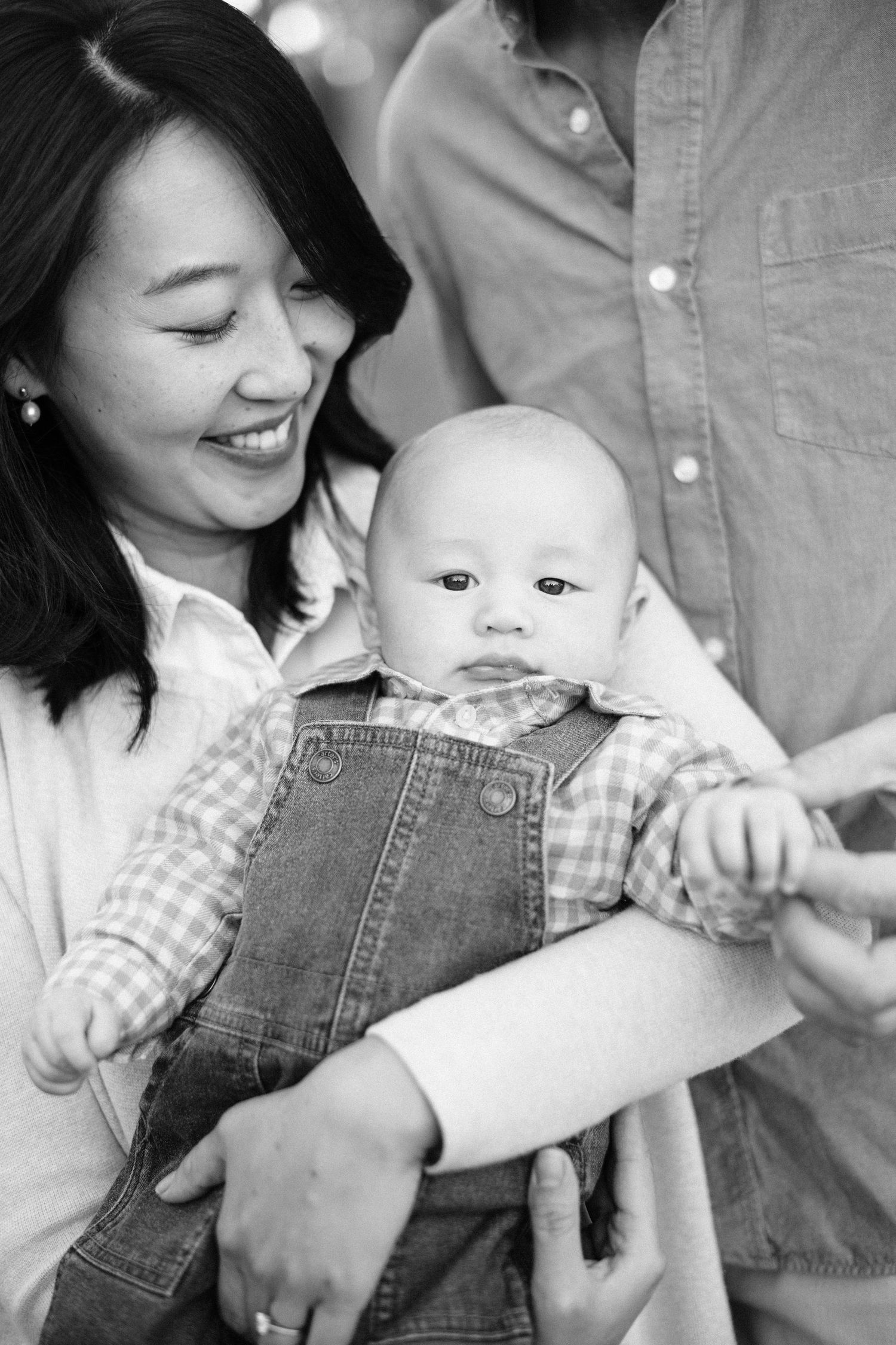 Black and white photo of a smiling Asian mother holding her baby in denim overalls, with father standing nearby.