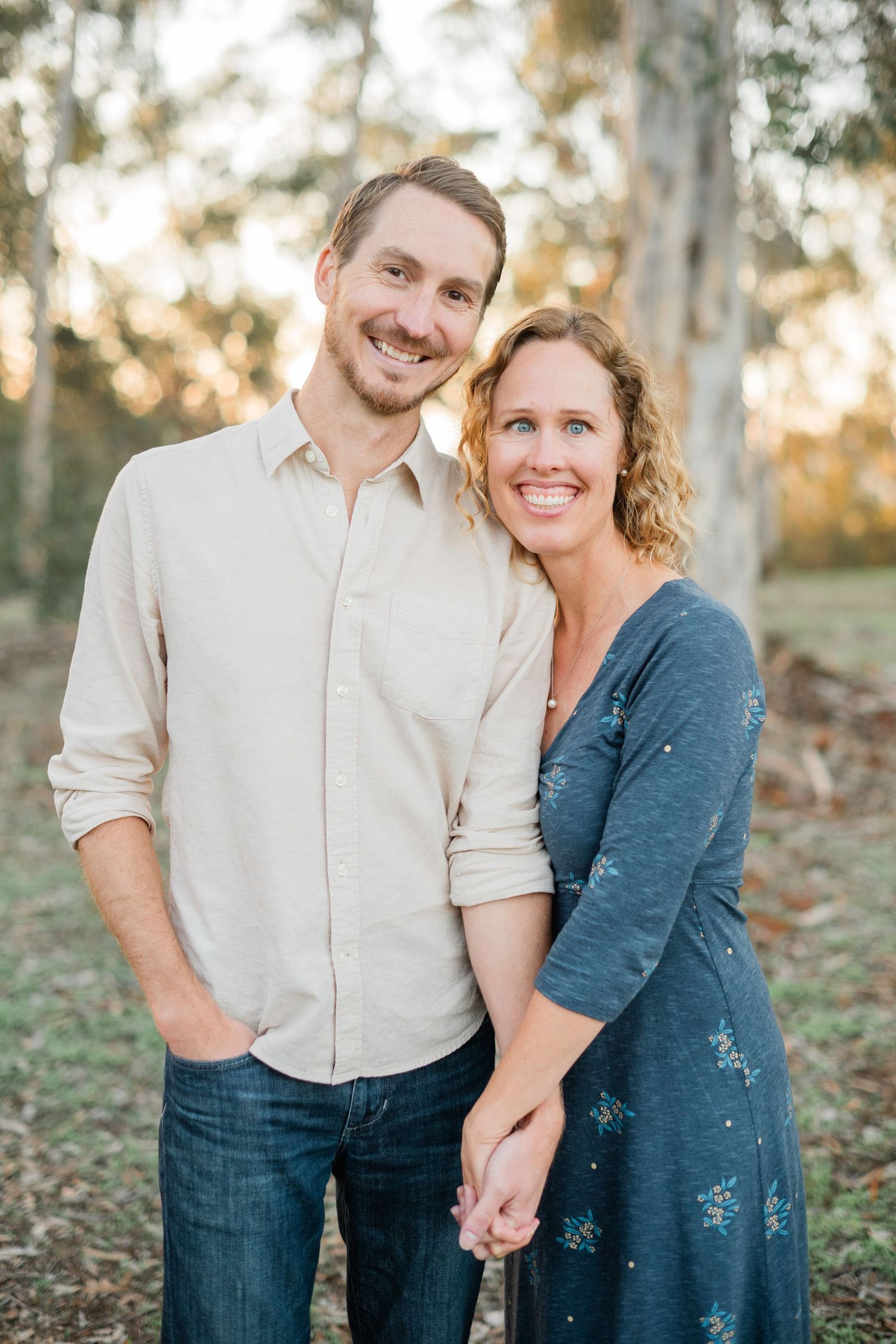 Smiling couple holding hands outdoors in a sunlit forest, man in beige shirt and jeans, woman in blue floral dress.