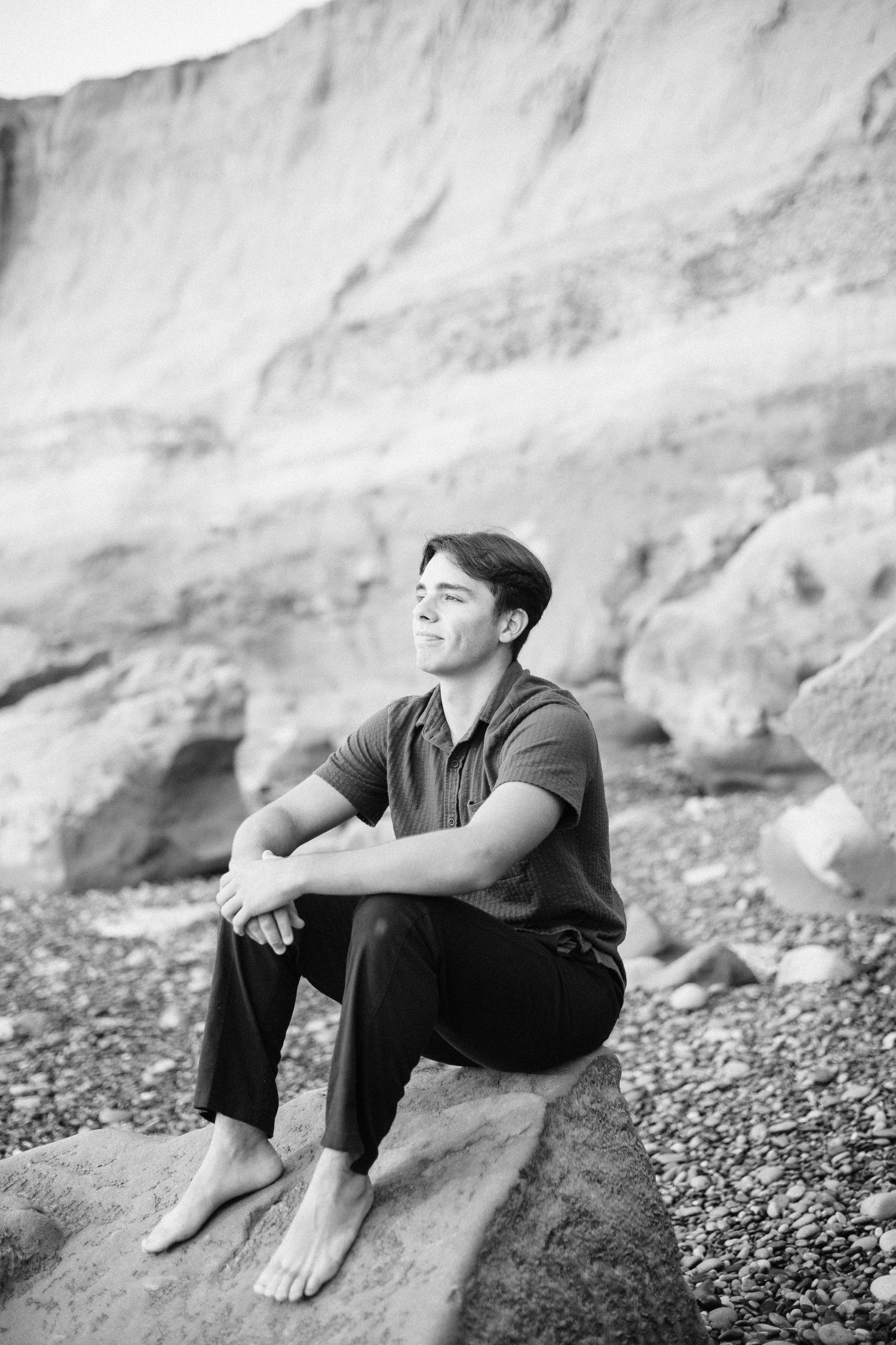 Black and white photo of a young man sitting barefoot on a rock at the beach, gazing upward.
