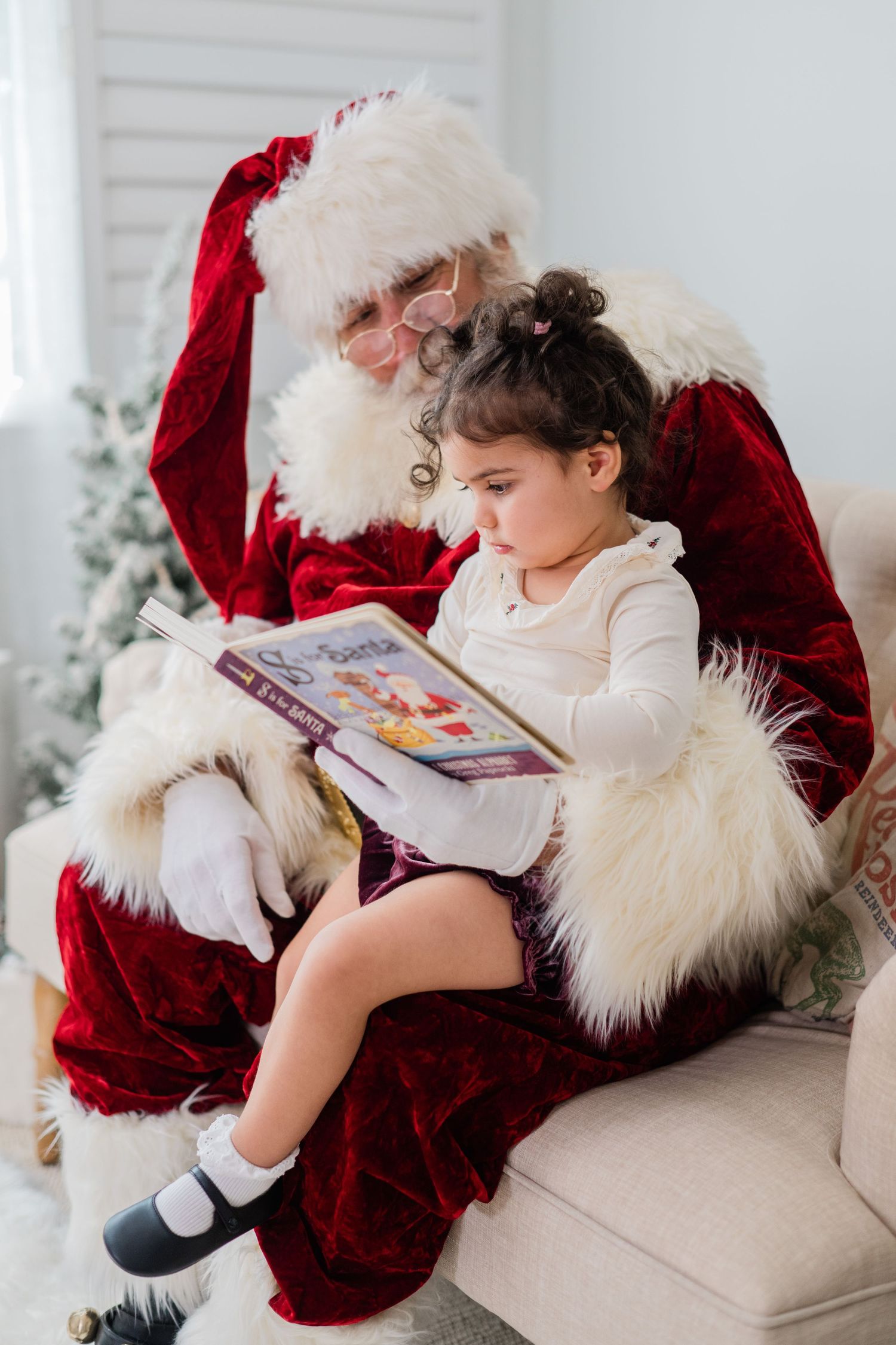 Santa Claus reads a colorful Christmas book with a curly-haired toddler girl sitting on his lap in a bright room.